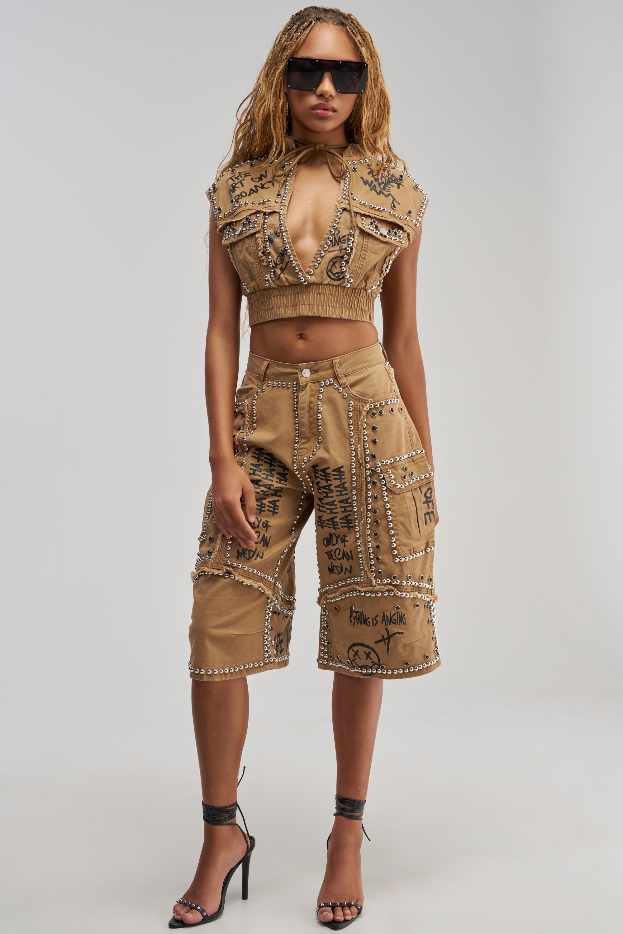 Wearing sunglasses, a woman styles the GET INTO THE RHYTHM BERMUDA SHORT—tan Bermuda shorts adorned with studs, patches, and bold black graffiti-style text—paired with a matching tan crop top vest and black high heels.