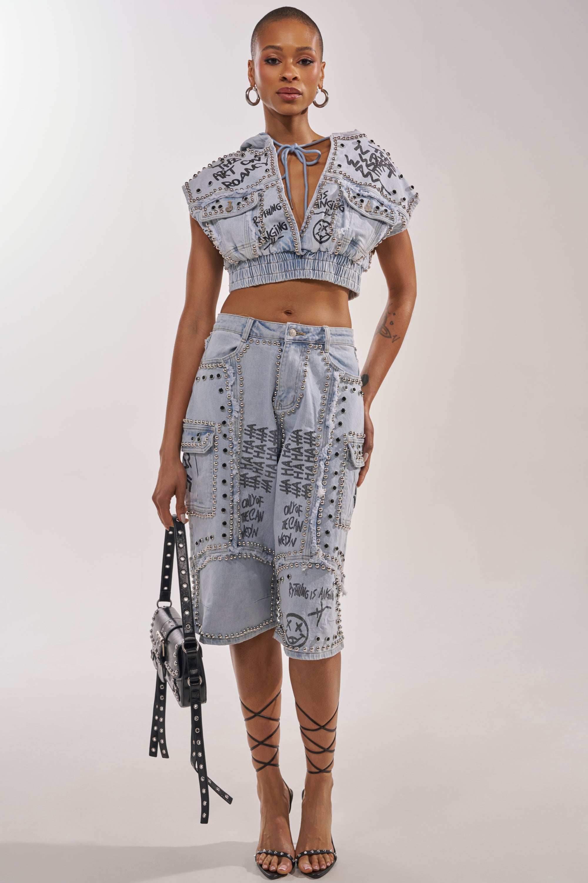 A woman wears the GET INTO THE RHYTHM BERMUDA SHORT, a light denim knee-length piece with black graffiti streetwear design, silver studs, cargo pockets, and details. She pairs it with a matching crop top and black studded accessories. 0