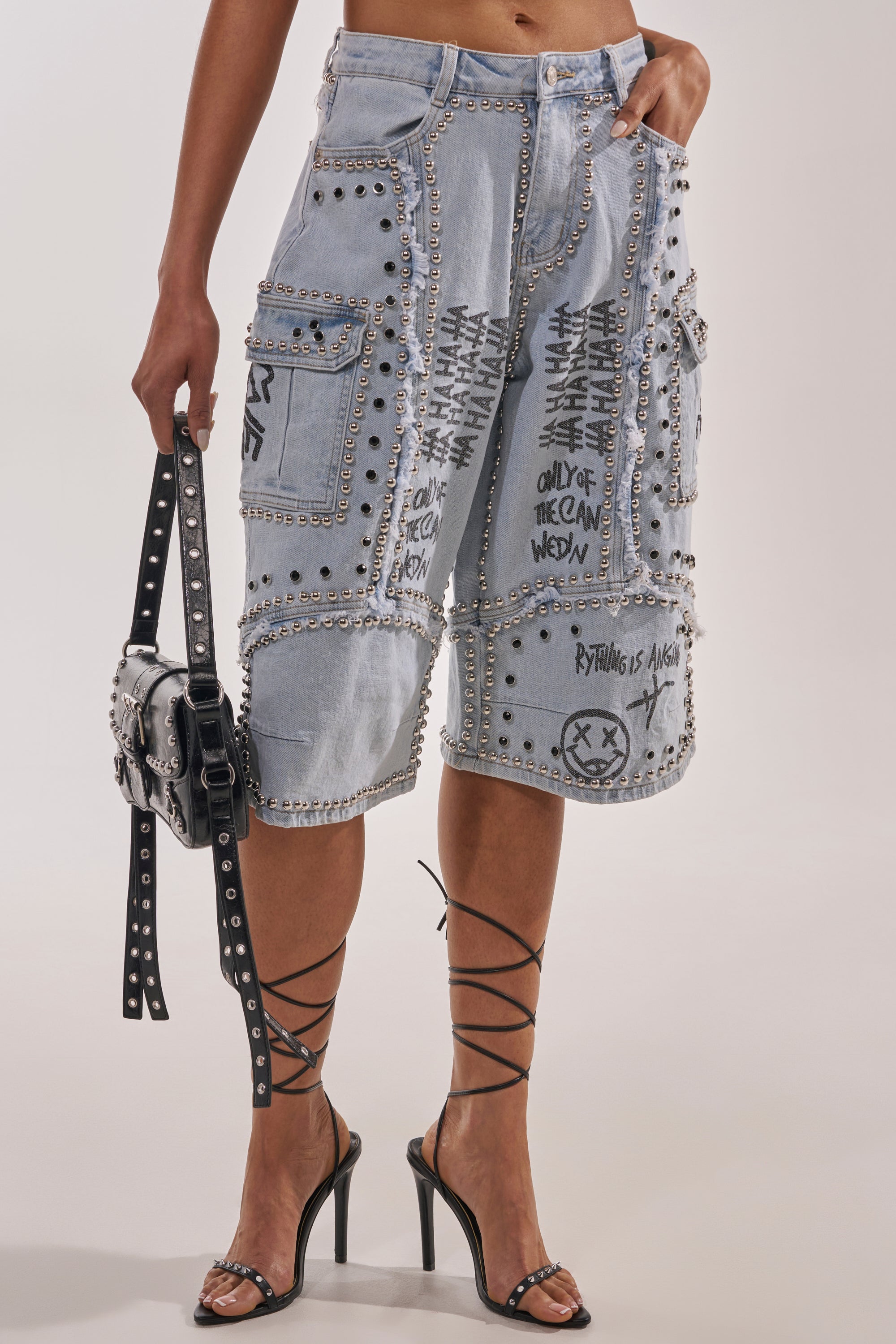 A person wears the GET INTO THE RHYTHM BERMUDA SHORT—light-wash denim with graffiti streetwear accents, metal studs, and cargo pockets—paired with black strappy stilettos and a studded handbag. Upper body not visible. 1