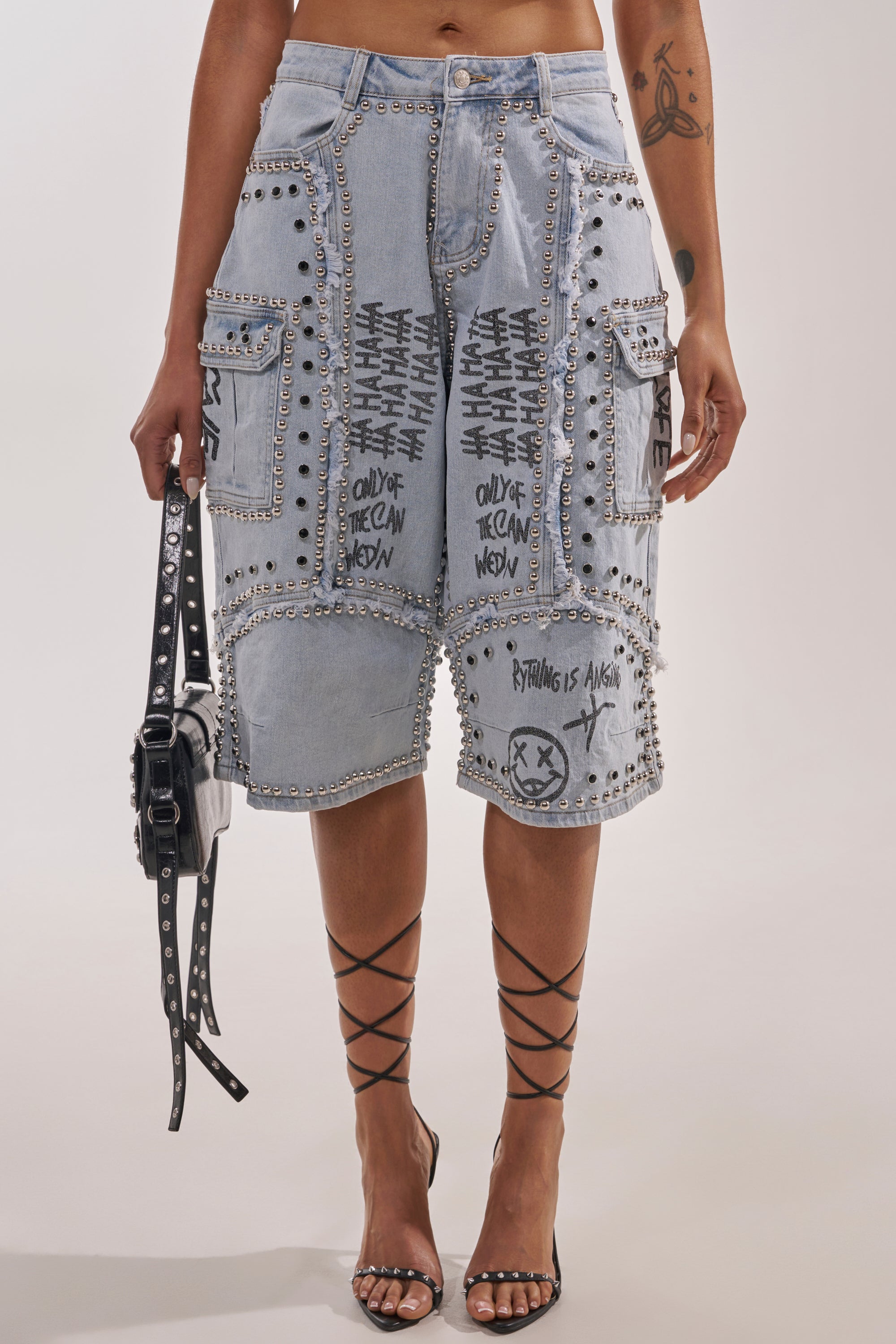 Wearing the GET INTO THE RHYTHM BERMUDA SHORT—light blue denim with graffiti graphics, studs, and patches—paired with strappy heels, a studded bag, and showing off an arm tattoo for a bold, expressive streetwear look. 2