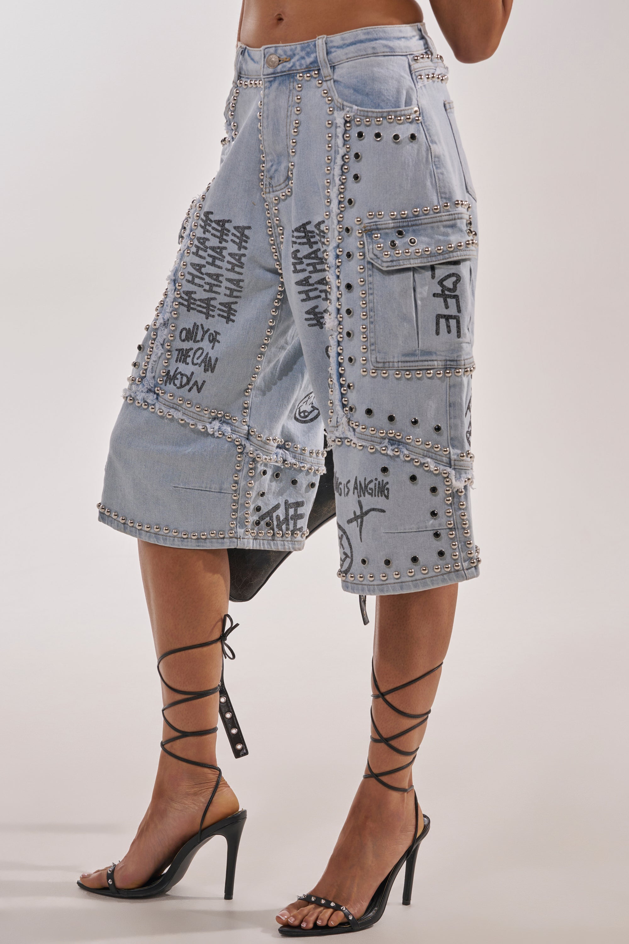 A person wearing the GET INTO THE RHYTHM BERMUDA SHORT—knee-length, light-wash denim with silver studs, black graffiti text, large cargo pockets—and strappy high-heeled sandals. The torso is cropped out of the image. 4