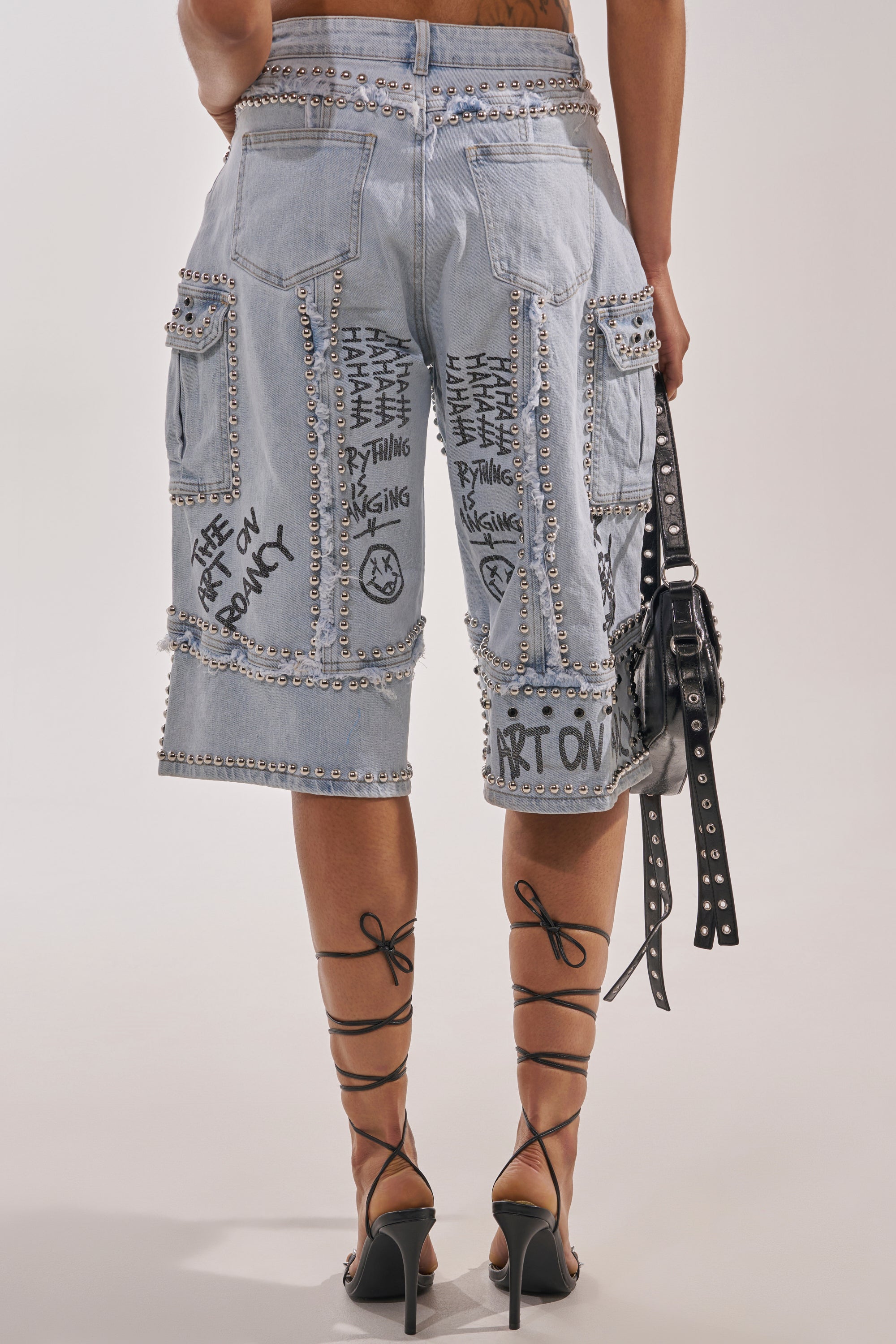 A person wearing the GET INTO THE RHYTHM BERMUDA SHORT—light blue, knee-length denim with patches, studs, cargo pockets, and black graffiti streetwear text—paired with strappy heels and a studded black handbag. Shot from behind. 5