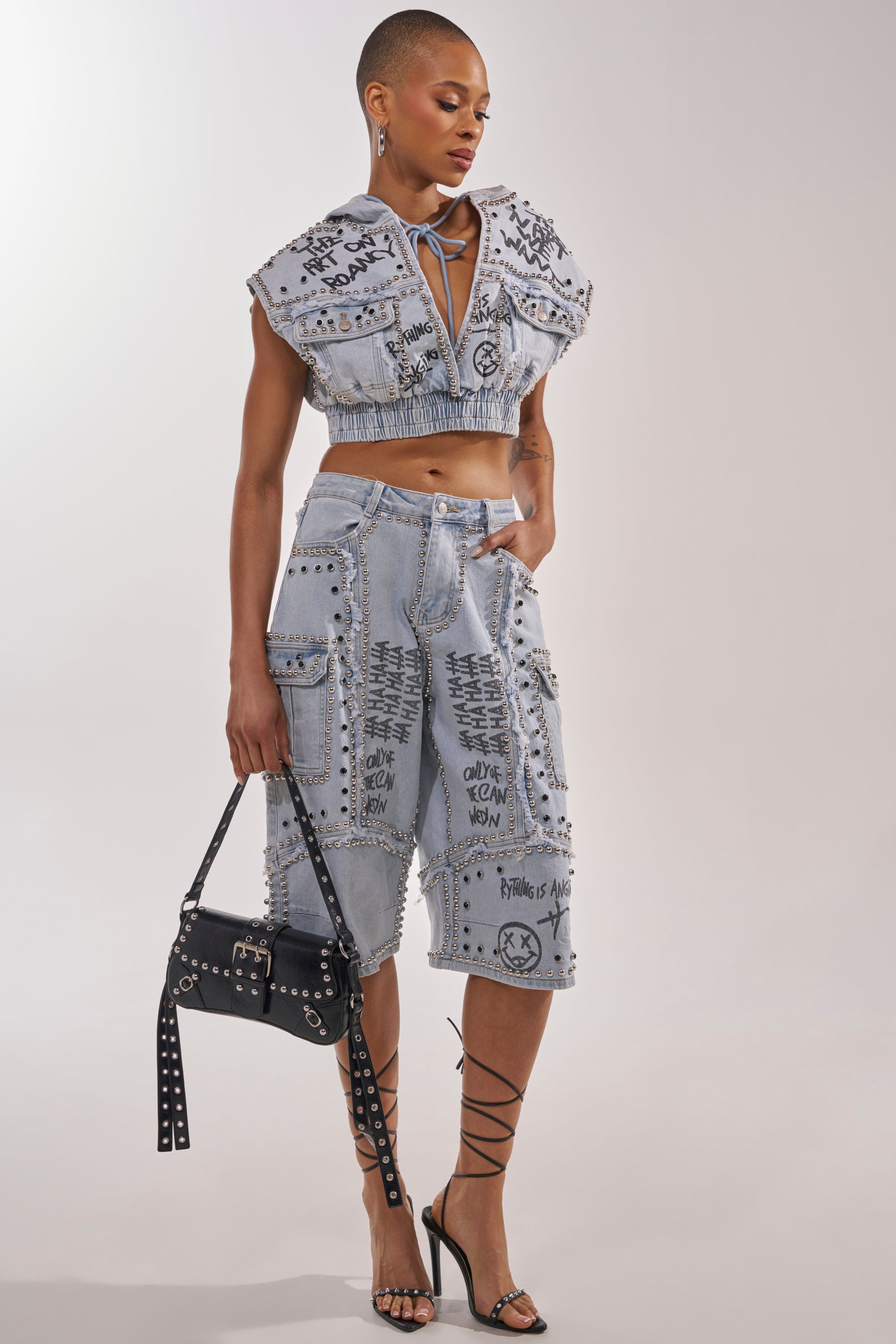 A woman styles the GET INTO THE RHYTHM BERMUDA SHORT, featuring light denim and graffiti details with cargo pockets, paired with a matching cropped top, strappy heels, and a black studded handbag. 6
