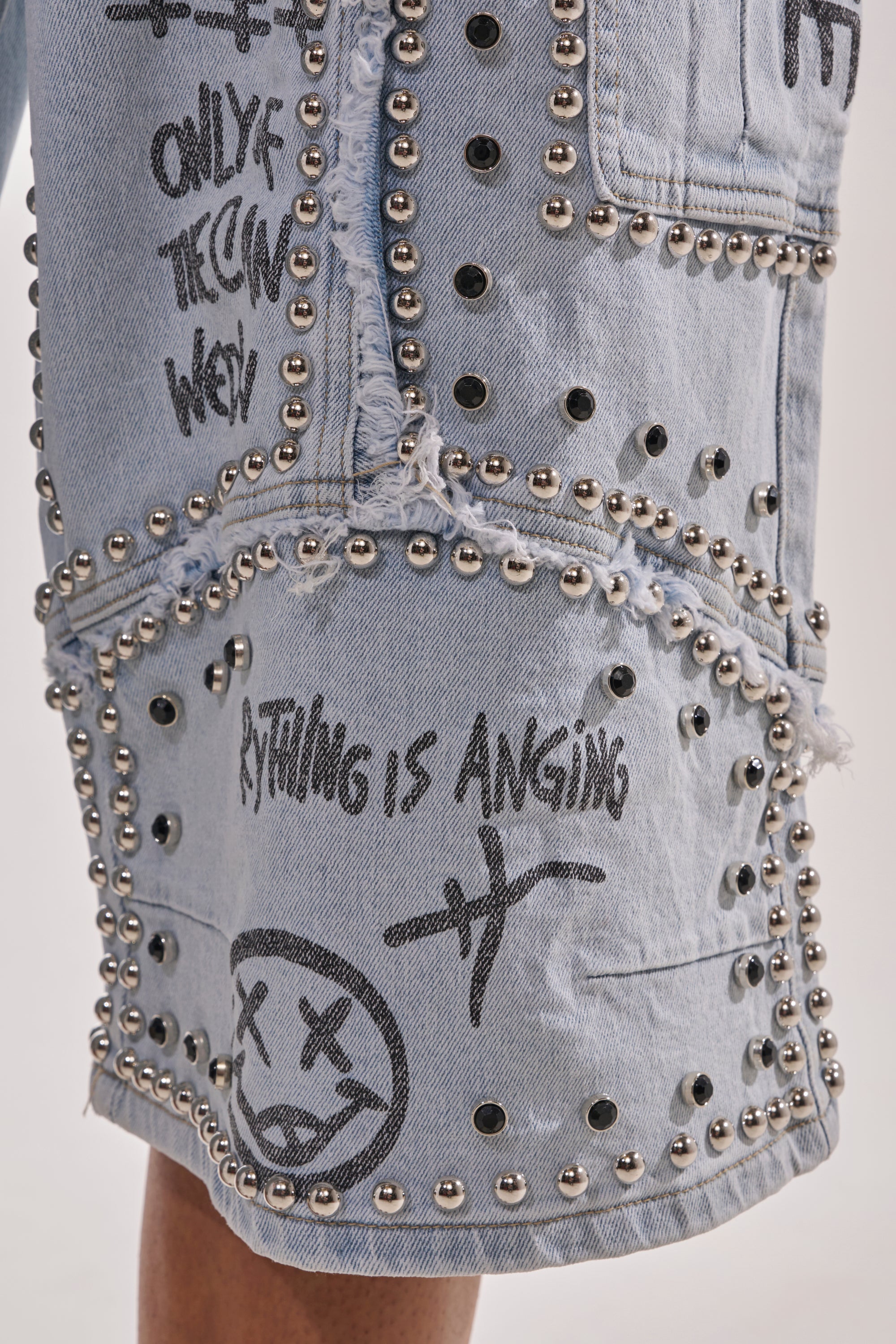Close-up of the GET INTO THE RHYTHM BERMUDA SHORT: streetwear denim featuring silver studs, bold graffiti-style graphics, edgy phrases, and a sketched face with X eyes and a wavy mouth. 7