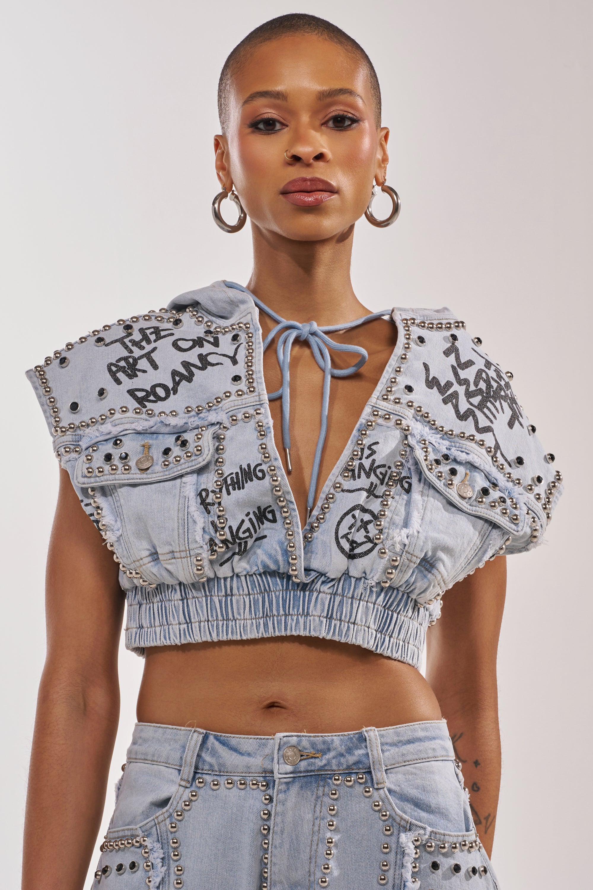 A woman rocks the GET INTO THE RHYTHM CROP HOODIE—sleeveless denim with metal studs and graffiti graphics—plus matching pants, hoop earrings, a shaved head, and a confident attitude for bold street-chic style. 1