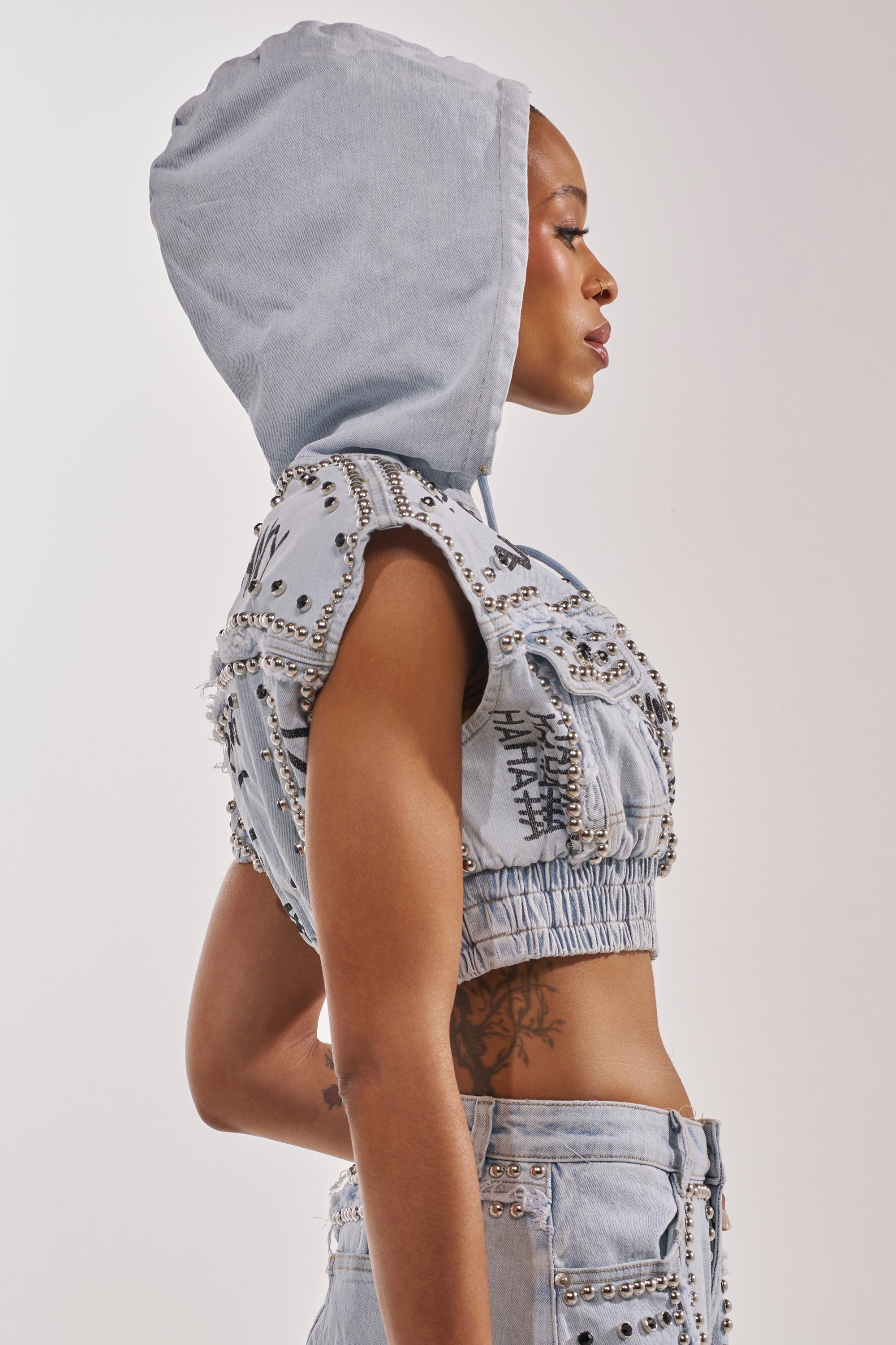 Wearing the GET INTO THE RHYTHM CROP HOODIE, this person poses in profile with matching studded jeans, showcasing tattoos and street-chic style against a plain background. 2