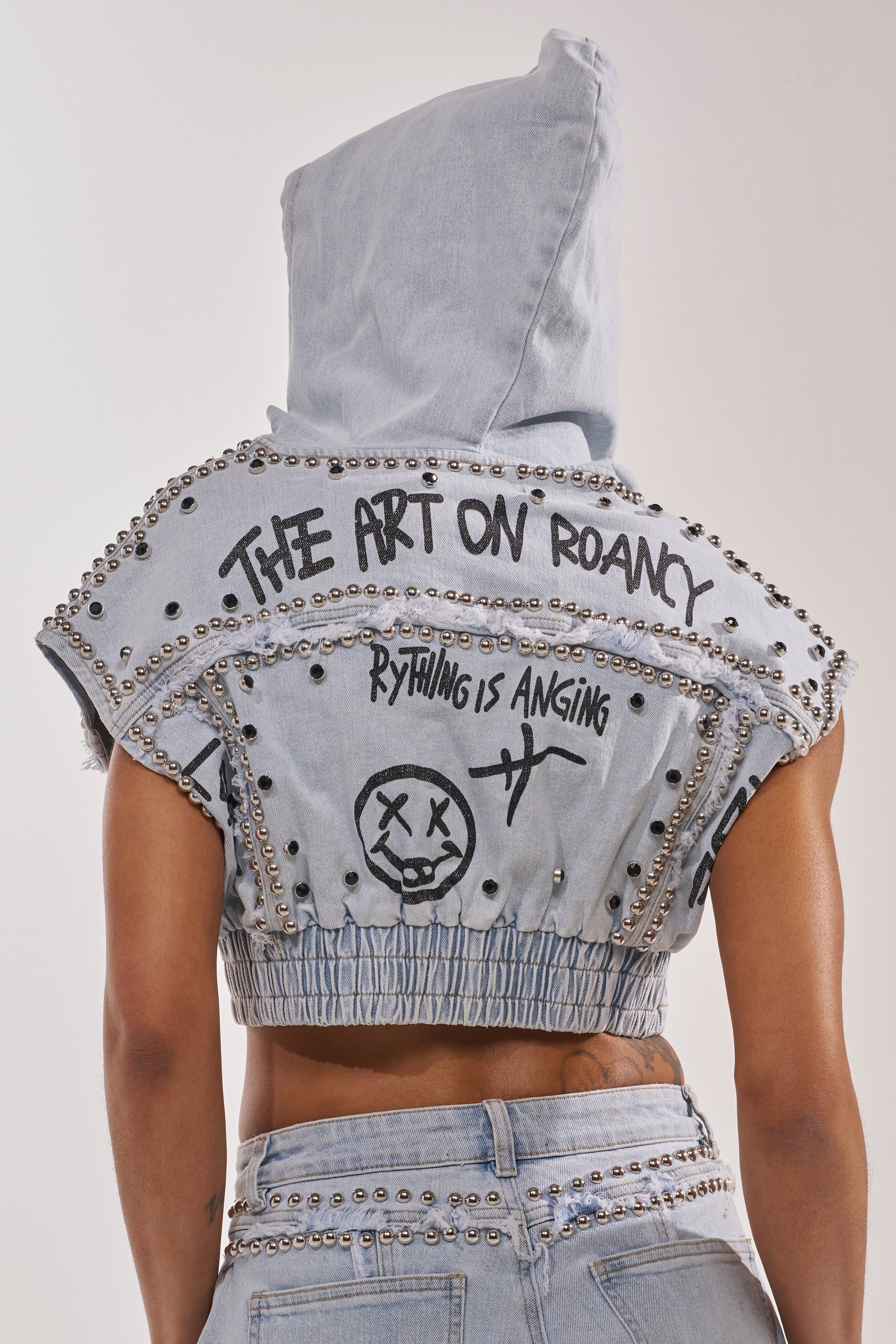 A person wears the GET INTO THE RHYTHM CROP HOODIE, a sleeveless denim piece with metal studs and graffiti-style graphics—"THE ART ON ROANCY," "RYTHING IS ANGING," and a doodled face with symbols—for effortless street-chic layering. 3