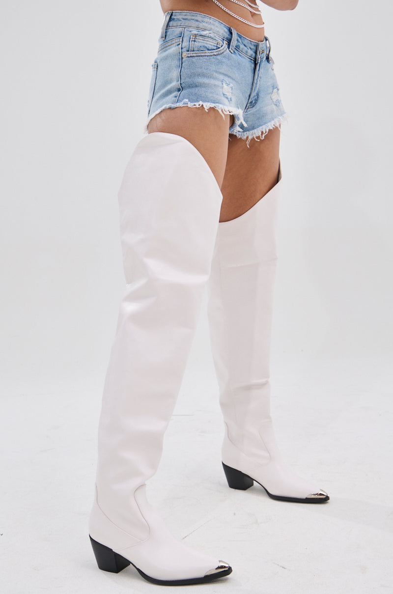 The image shows the lower half of a person wearing distressed denim shorts and the AZALEA WANG RIA OVER THE THIGH WESTERN BOOT IN WHITE. These boots feature a faux leather thigh-high shaft with black heels, set against a plain white background.