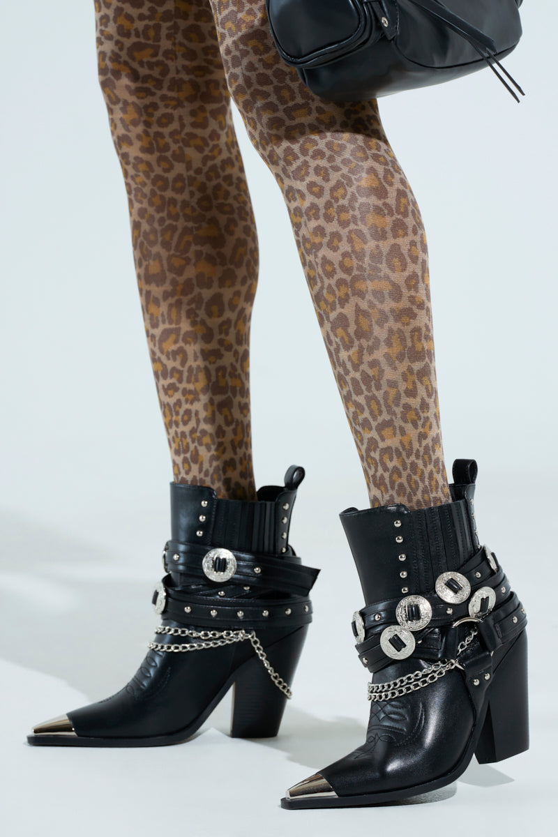 The AZALEA WANG RIGGINS black booties, featuring silver studs, buckles, and chains, stylishly pair with leopard print leggings. Crafted from vegan leather with a pointed-toe design and metallic accents, these Western-style ankle boots are completed with a black purse for a bold look.