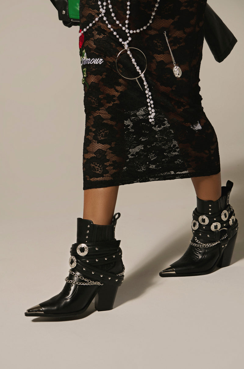 Wearing AZALEA WANG RIGGINS black silver hardware embellished Western Bootie with chunky high heels, paired with a black lace skirt adorned in floral embroidery and pearl accents, stands out against the plain light background.