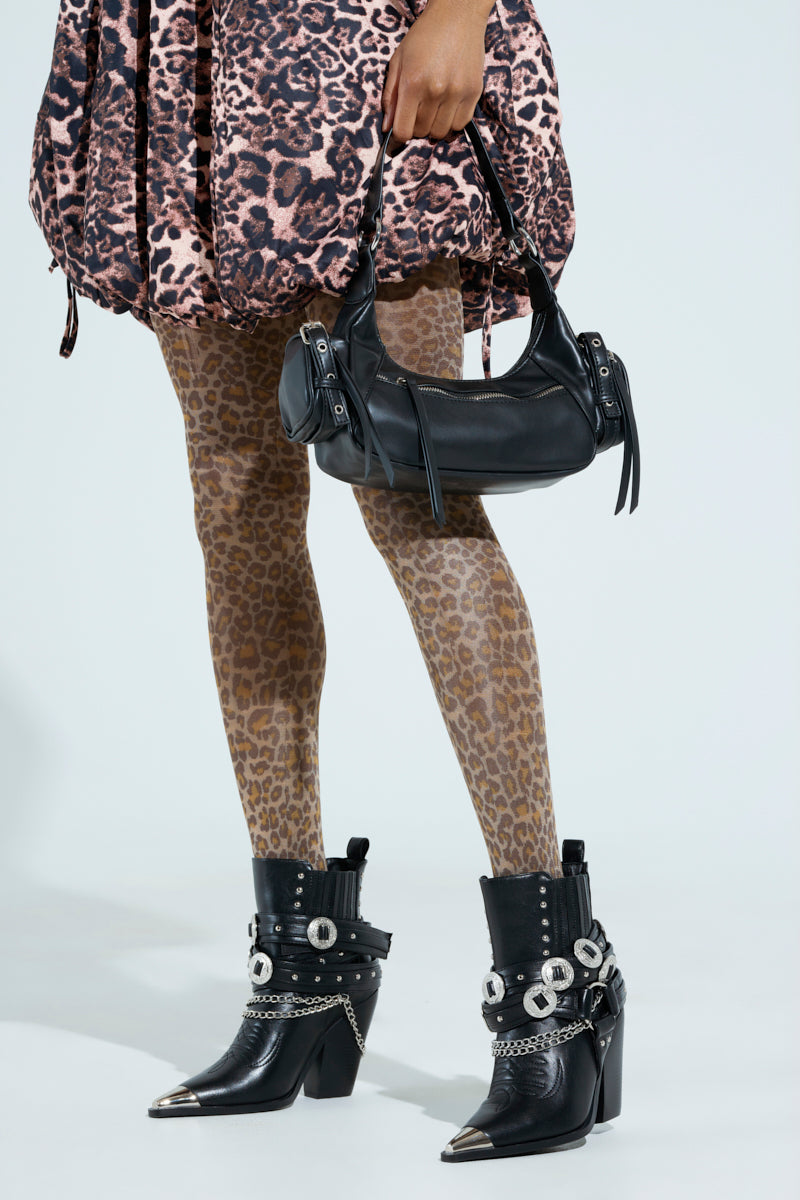 Dressed in a leopard print skirt and tights, someone holds a small black vegan leather handbag while wearing AZALEA WANG RIGGINS black heeled boots, embellished with silver chains and buckles. The background is plain white.