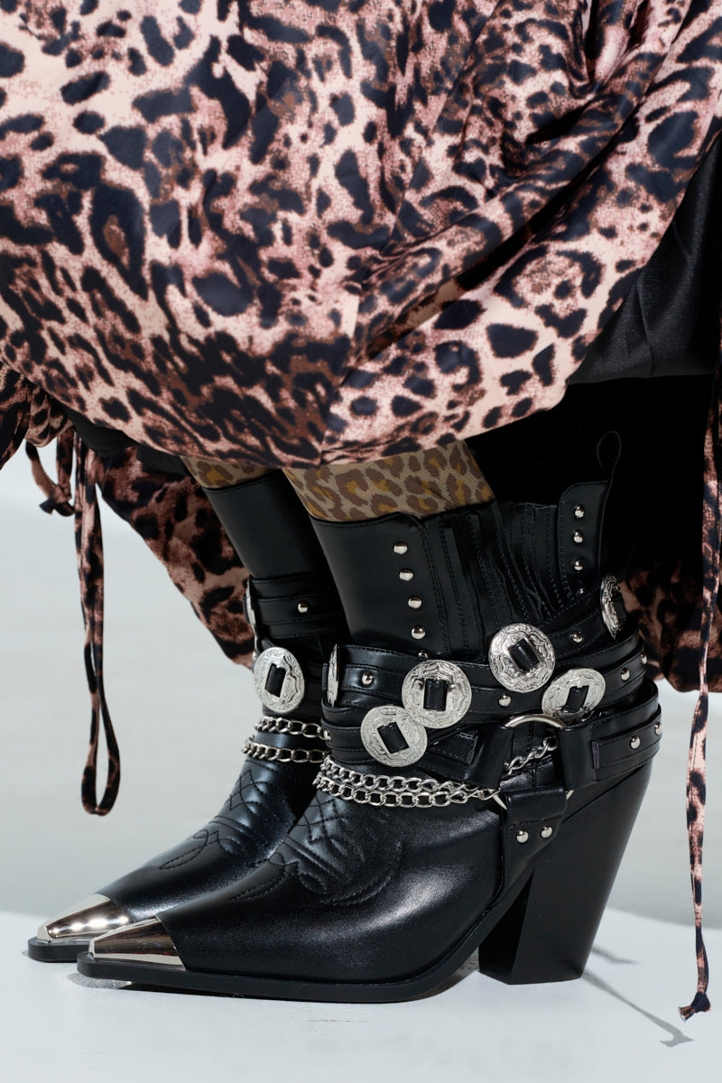 The AZALEA WANG RIGGINS black silver hardware embellished Western booties with high heels and pointed metal tips pair boldly with a leopard-print skirt.