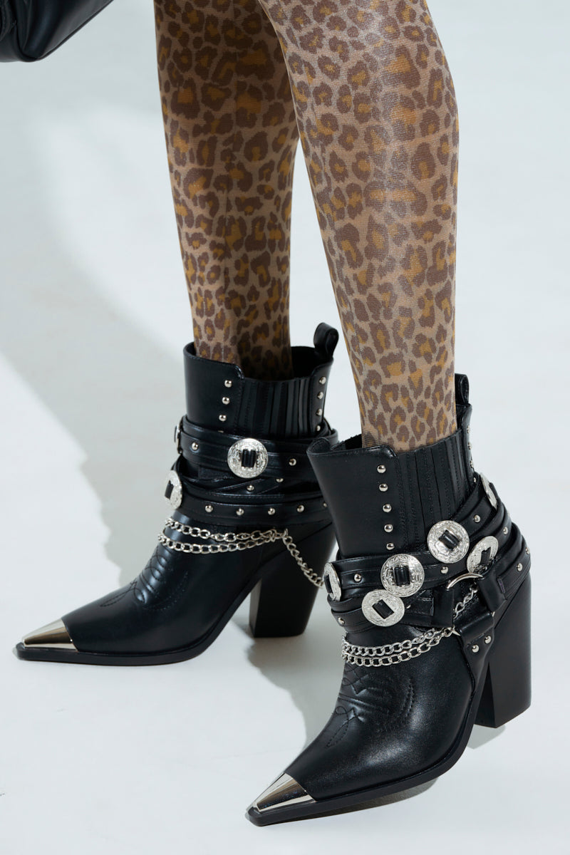 A person wearing leopard print leggings paired with AZALEA WANG RIGGINS black vegan leather ankle boots, adorned with silver buckles, studs, chain embellishments, high block heels, and metallic hardware toe caps.