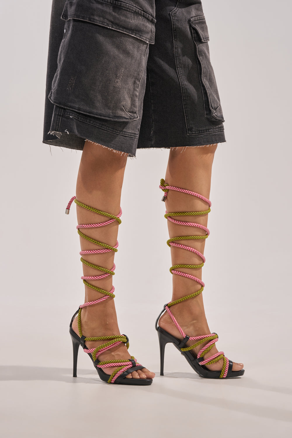 A person in black denim cargo shorts and eye-catching multicolor AZALEA WANG RIJN ROPE STILETTO LACE UP SANDALS, featuring long pink and yellow wraparound rope straps on chic stiletto heels. 1