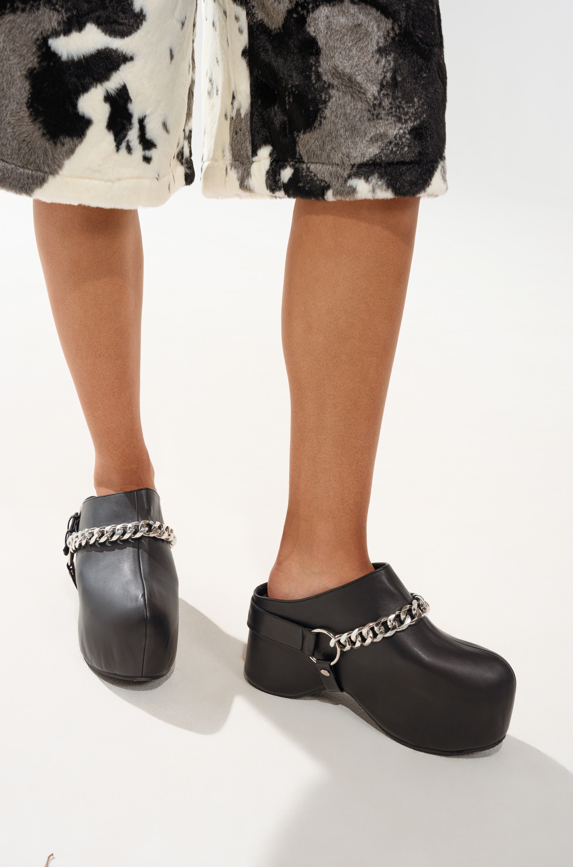 A person wearing the AZALEA WANG RILLMEY BLACK FLAT CLOG WITH METAL DETAIL, paired with a knee-length skirt with a black and white abstract pattern, stands against a white background.