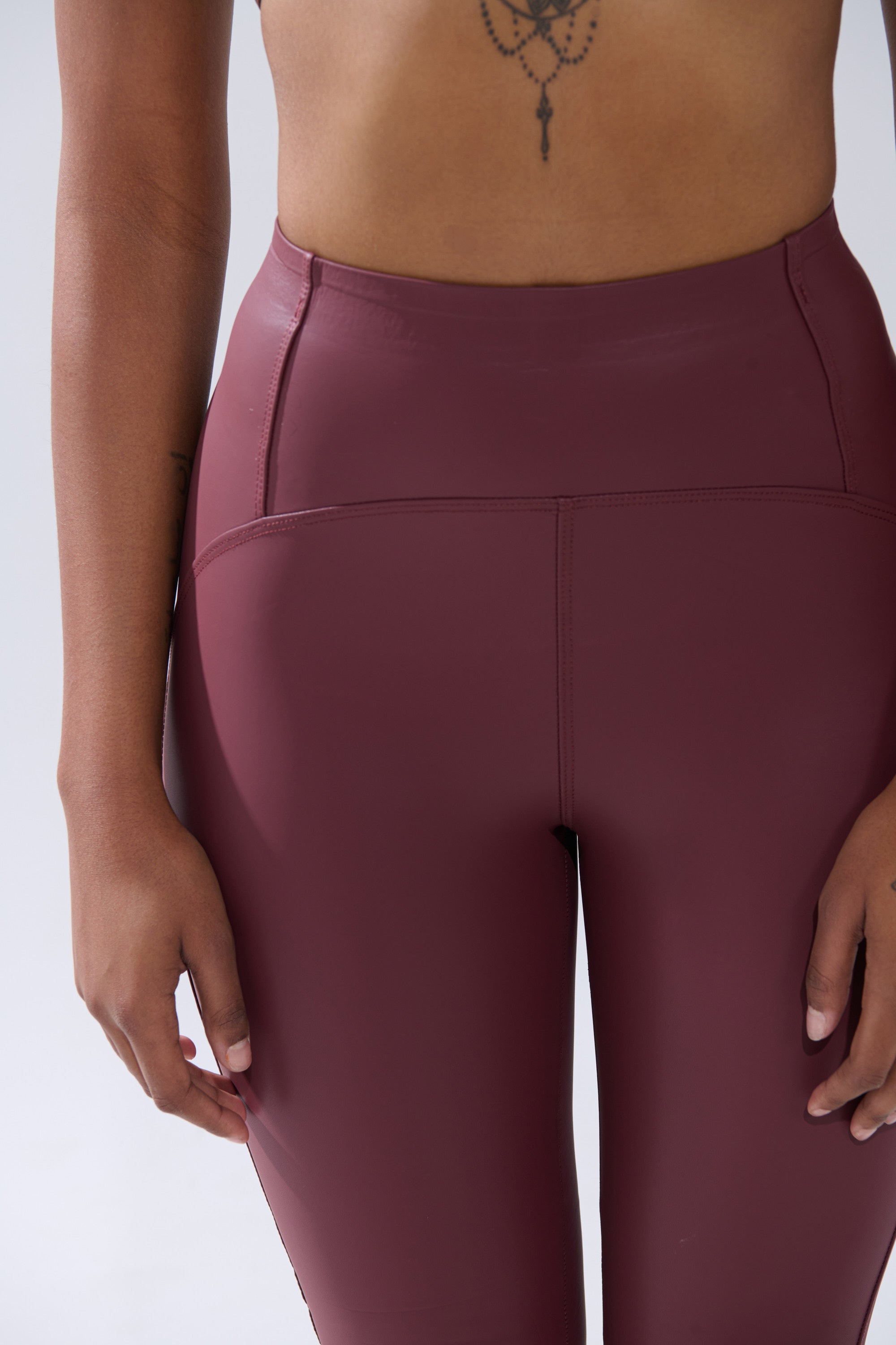Model wears RIO HIGH RISE LEGGING WITH 4 WAY STRETCH IN OXBLOOD, shown from waist to mid-thigh against a plain light background; hands relaxed at sides and part of a chest tattoo visible.