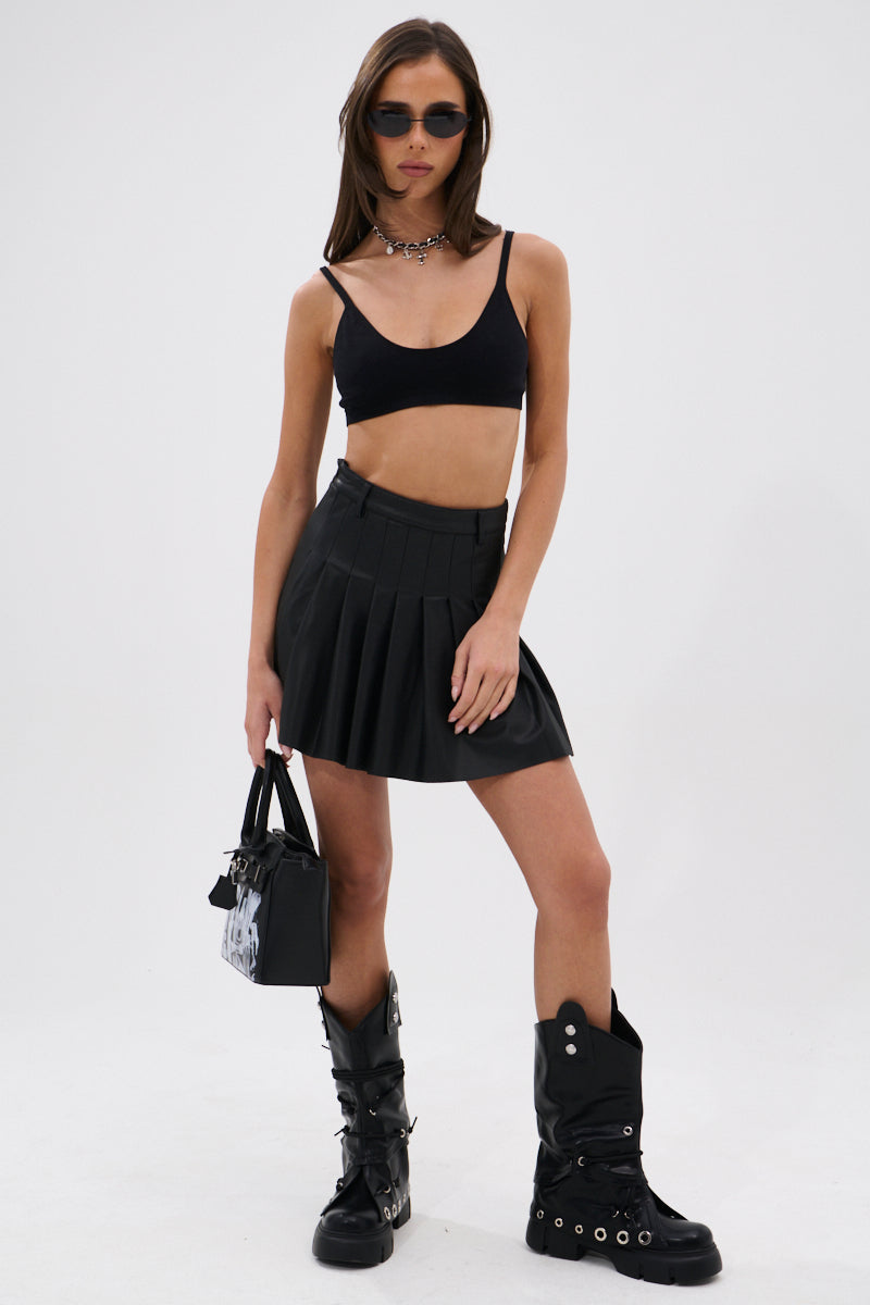 A woman stands confidently in sunglasses, a black crop top, chunky boots, and the RIO Pleated Faux Leather Mini Skirt with 4-Way Stretch. She holds a black handbag and wears a choker, posing against a plain white background.