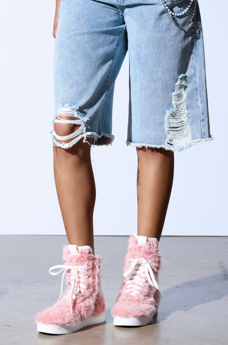 Wearing ripped denim shorts, a person stands on a concrete floor with their legs accentuated against a light gray wall by the AZALEA WANG RIORDAN PINK FUR HIGHTOP SNEAKERS, featuring bold faux fur uppers and white laces.
