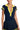 A woman is wearing the ALWAYS LUCKY RIBBED COLLARED BODYSUIT from AKIRA, which is navy blue with a yellow collar and a yellow button placket. The schoolgirl-inspired style includes yellow stripes on the sleeves. She complements it with a matching navy blue skirt featuring yellow trim at the hem, and accessorizes with large hoop earrings.