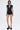 A woman stands facing forward in a short, fitted LAUREN SEAMLESS ROMPER WITH LACE featuring a deep V-neck and short sleeves. She wears glasses, black platform heels, and has straight shoulder-length hair against a plain white background.
