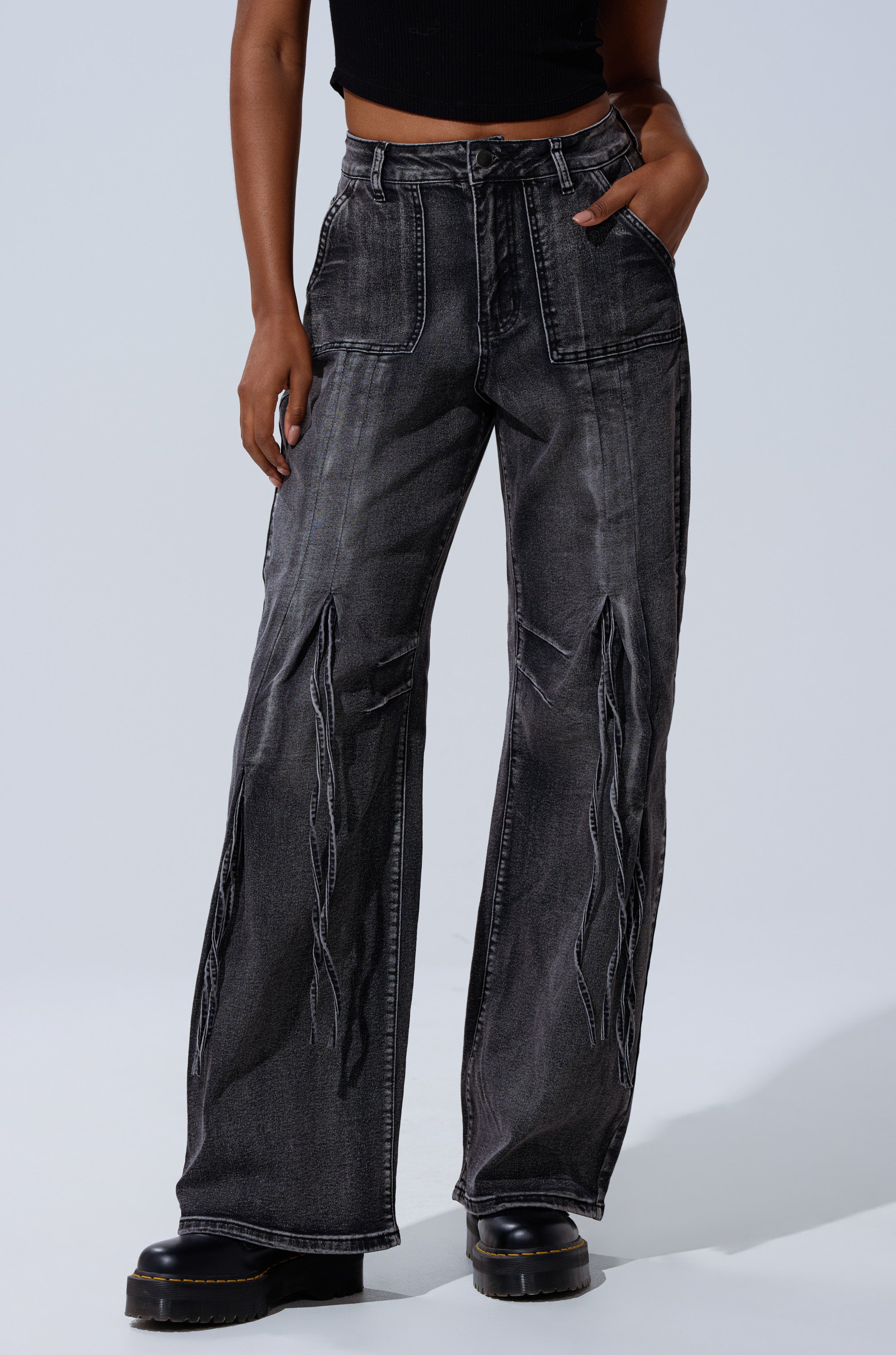 Someone wearing the TELL ME ABOUT IT TIE UP DENIM jeans, which feature high-rise fit, distressed seams, large front pockets, styled with a black sleeveless crop top and black platform shoes.
