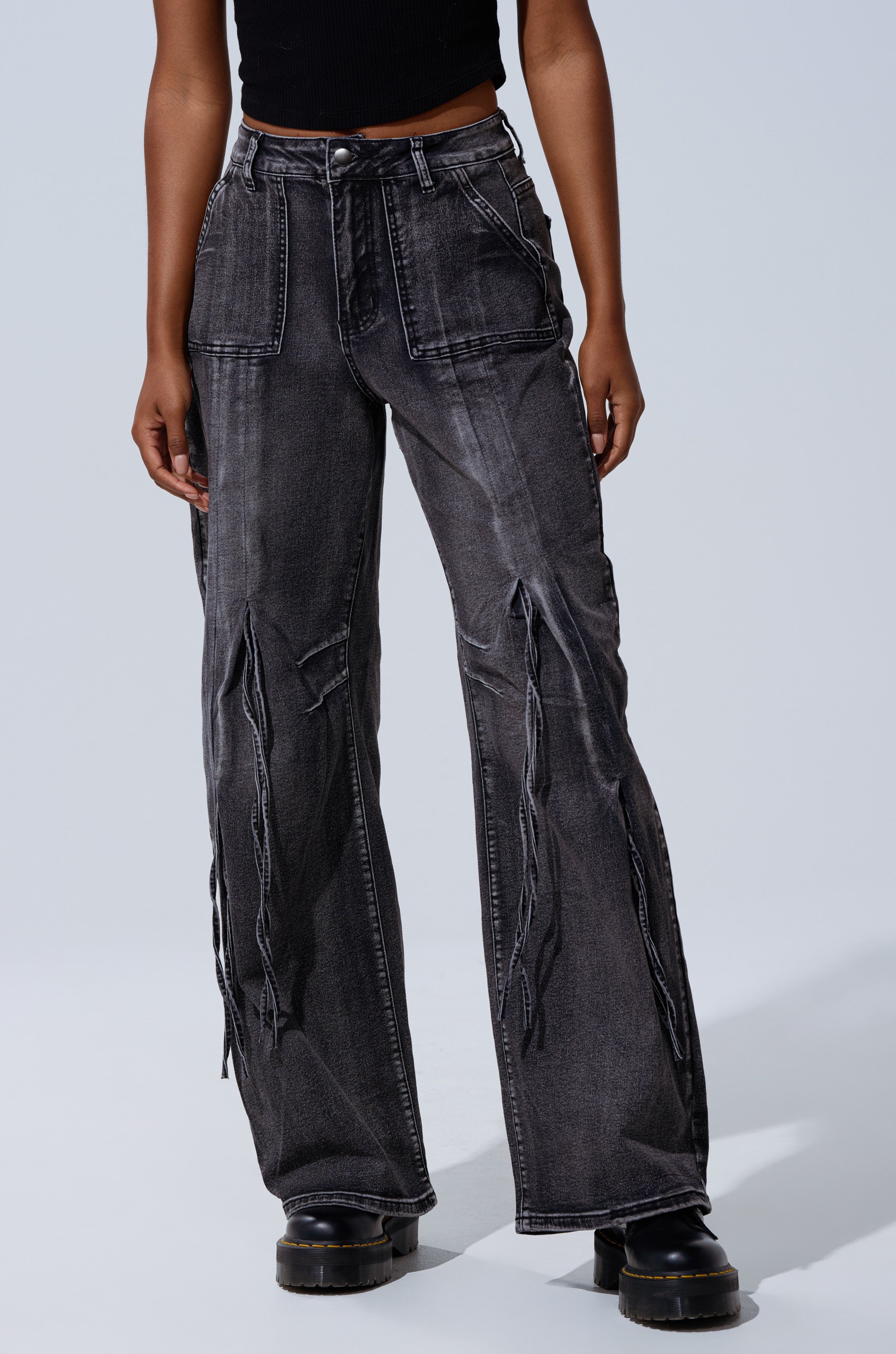 Person wearing the TELL ME ABOUT IT TIE UP DENIM: high-rise, wide-leg jeans with exposed seams and distressing, paired with a black sleeveless top and platform shoes, standing against a plain light background.