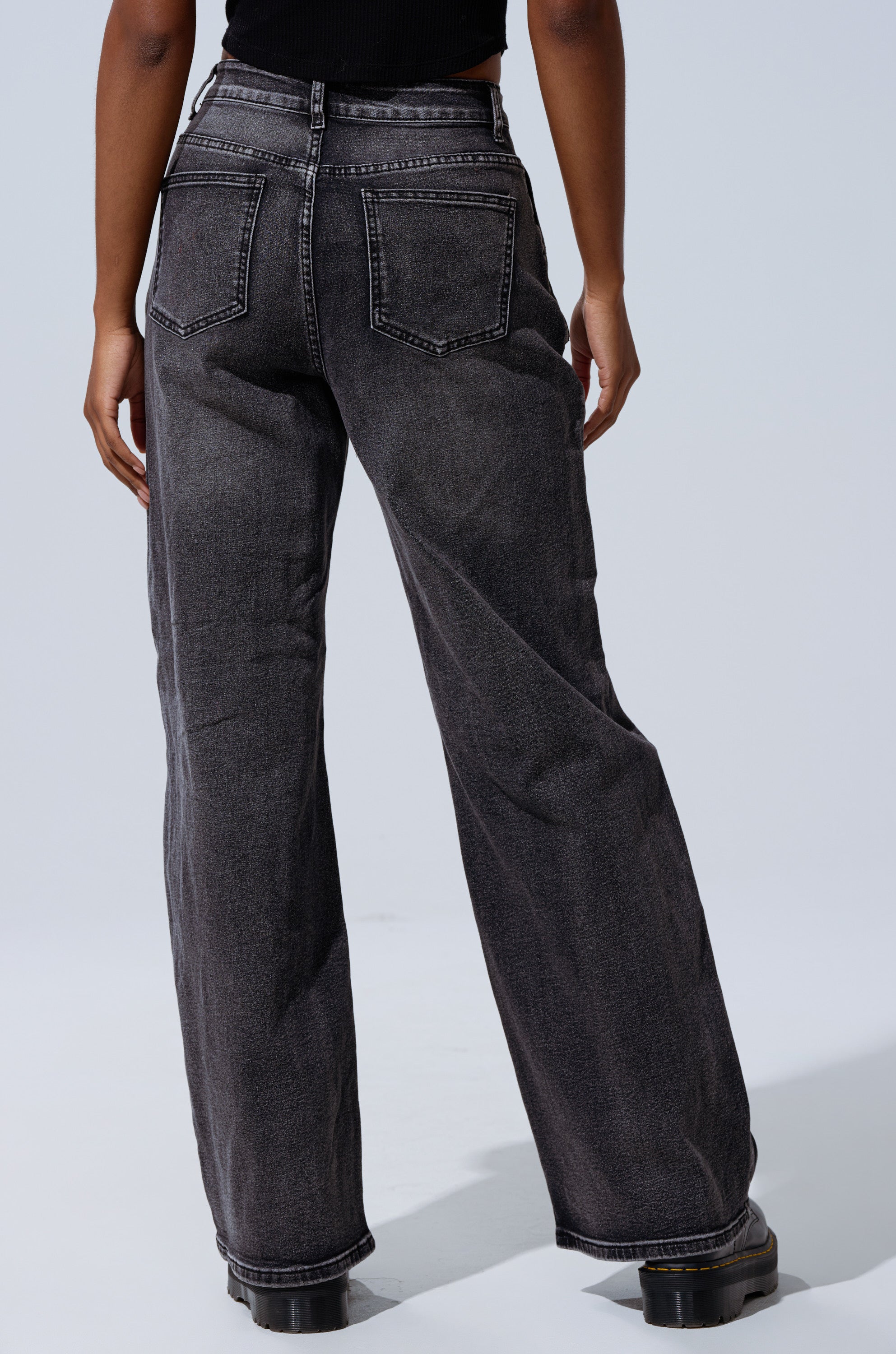 Someone faces away from the camera against a plain light background, wearing TELL ME ABOUT IT TIE UP DENIM high-rise wide-leg jeans in faded black and black shoes.