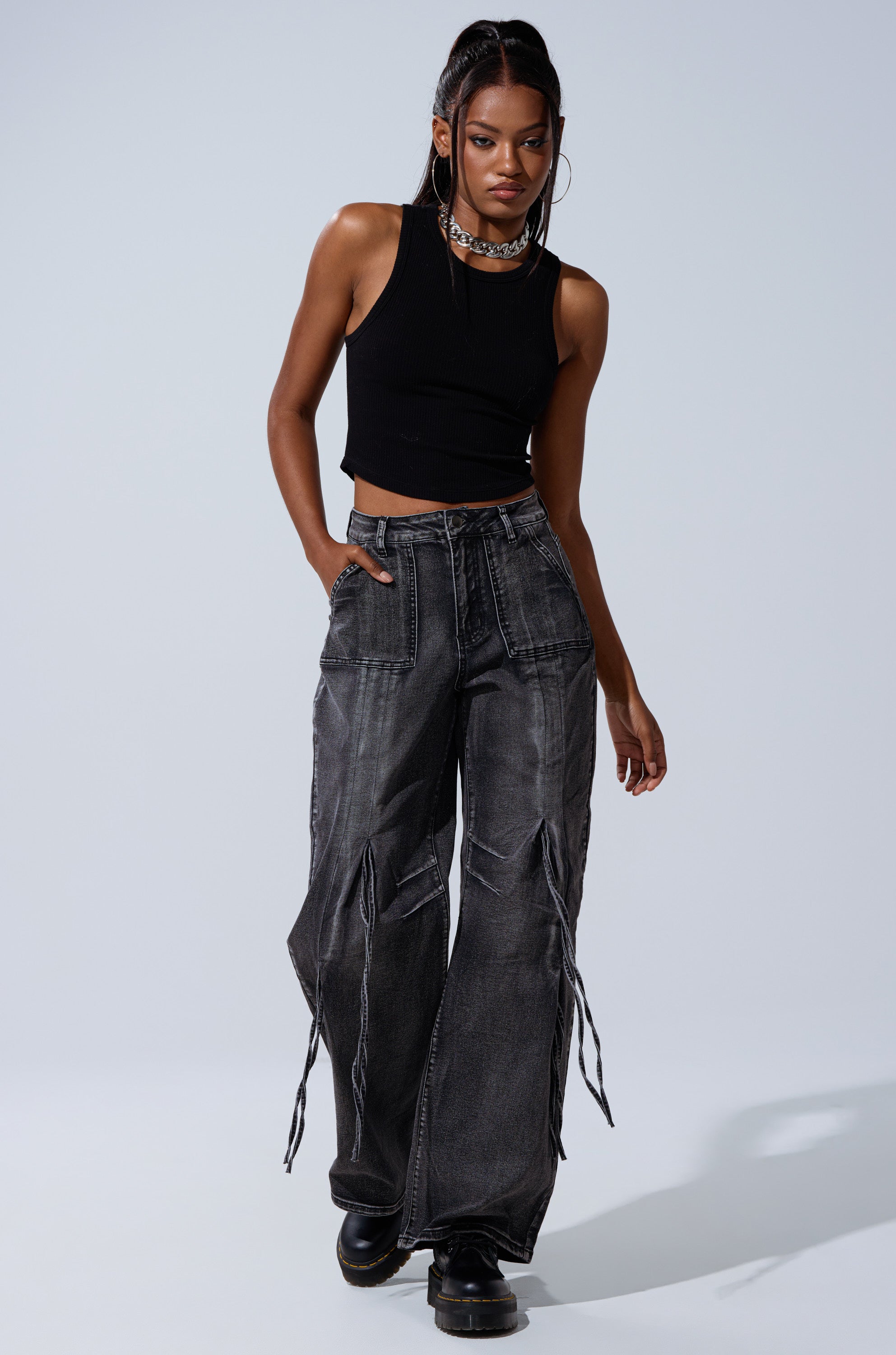 A woman with long dark hair poses confidently against a plain light background in the TELL ME ABOUT IT TIE UP DENIM—high-rise wide-leg jeans with bold white stitching—paired with a sleeveless black crop top and black platform shoes.
