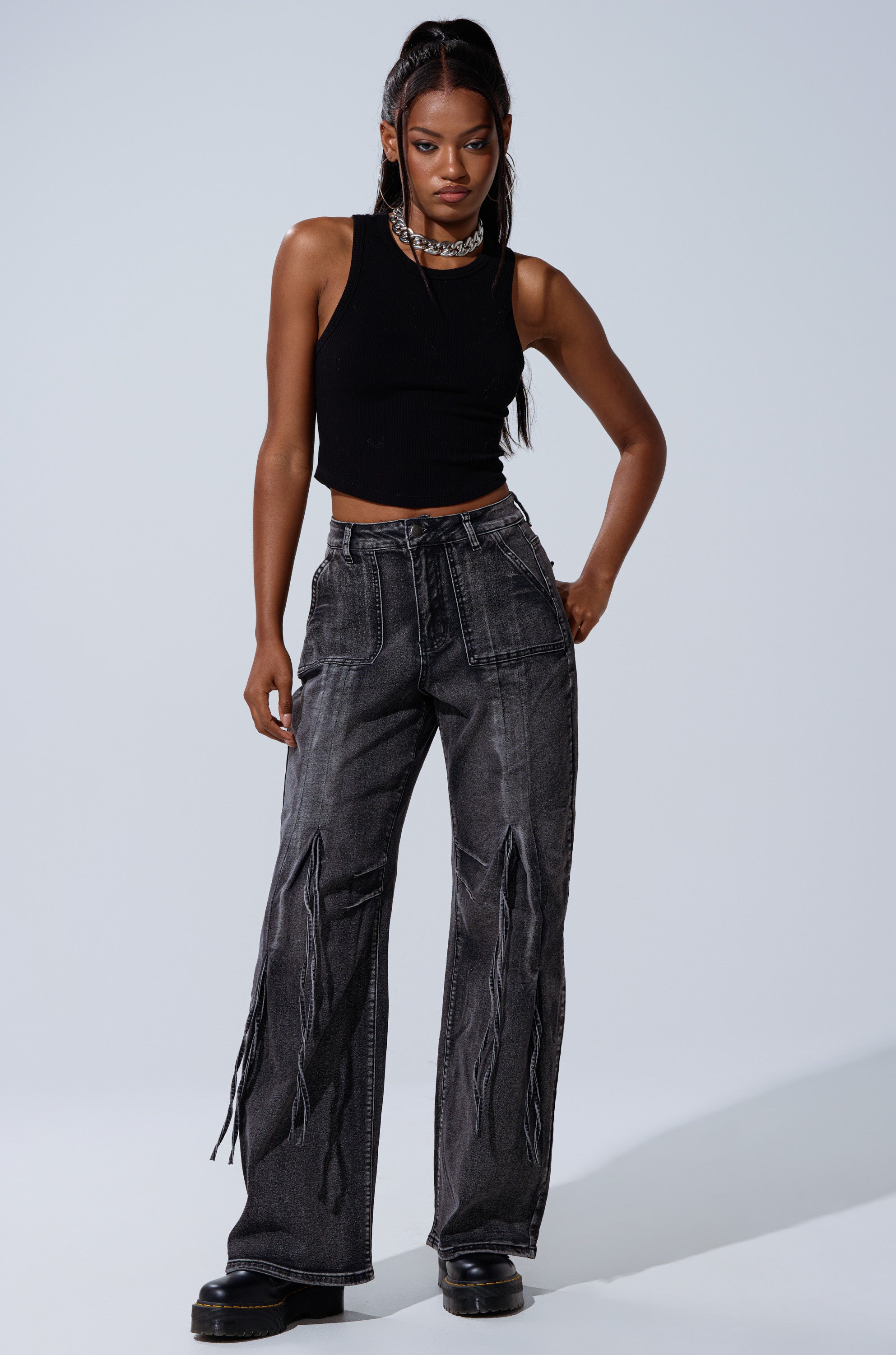 A woman with long dark hair wears the TELL ME ABOUT IT TIE UP DENIM jeans, a black sleeveless crop top, platform shoes, and layered necklaces, standing against a plain light background.