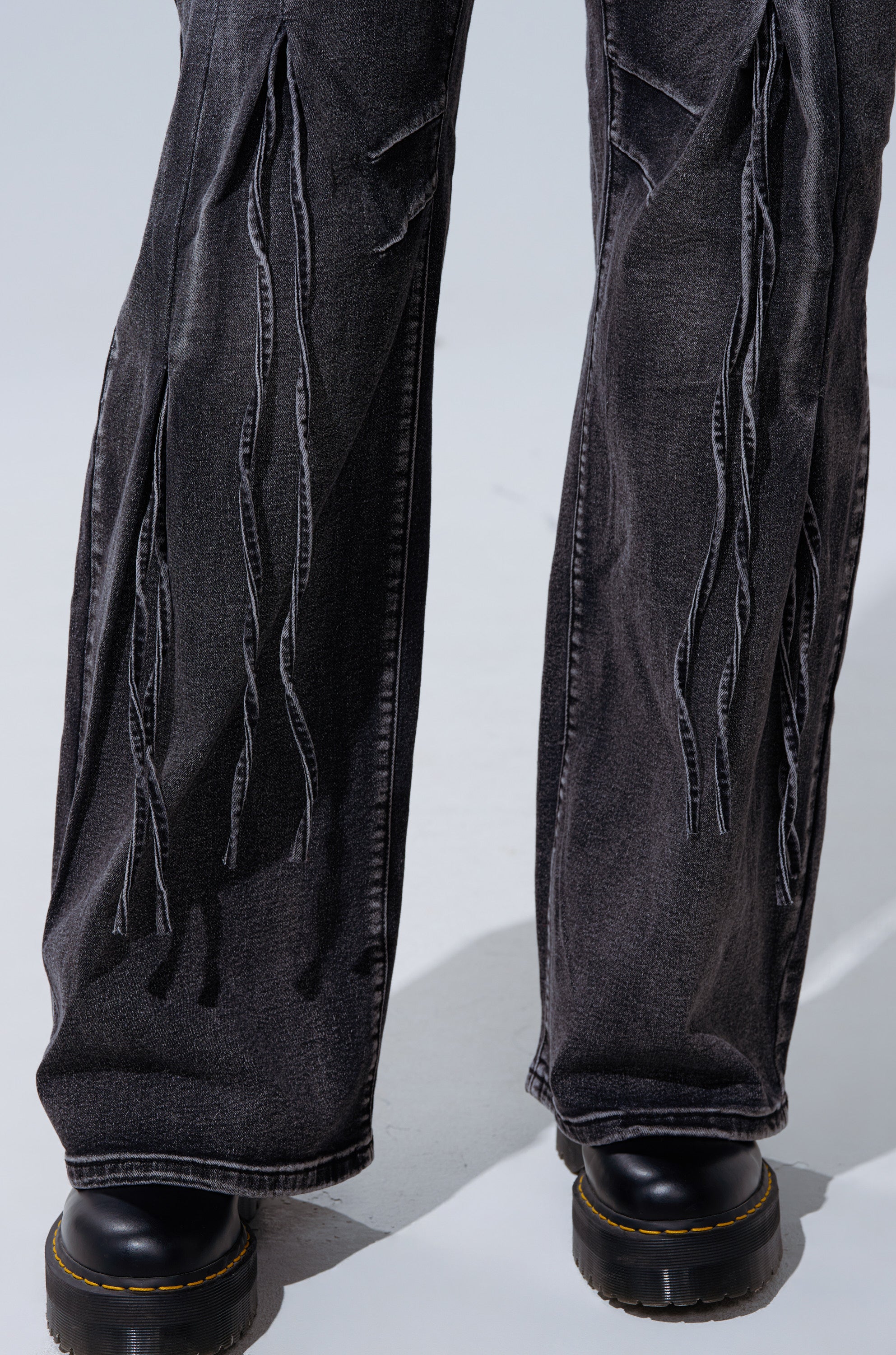 Close-up of a person wearing the TELL ME ABOUT IT TIE UP DENIM—black high-rise jeans with frayed vertical strips—paired with black chunky shoes with yellow stitching, standing on a light-grey surface.
