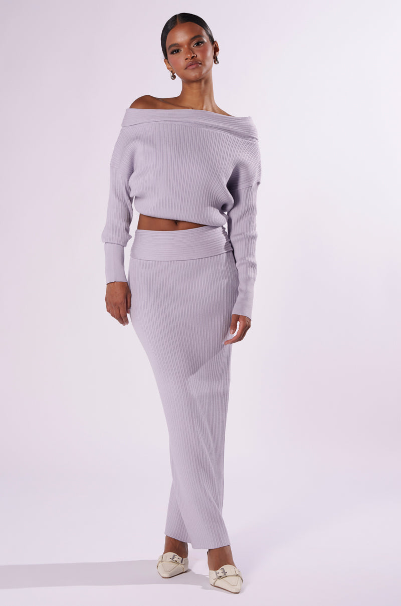 A person stands against a light grey background wearing the ribbed, light lavender CAN'T LOSE SWEATER KNIT MIDI SKIRT paired with an off-the-shoulder sweater. The high-rise bodycon skirt accentuates their silhouette. They have their hair pulled back and are dressed in white loafers, with one hand resting by their side in a relaxed posture. 4
