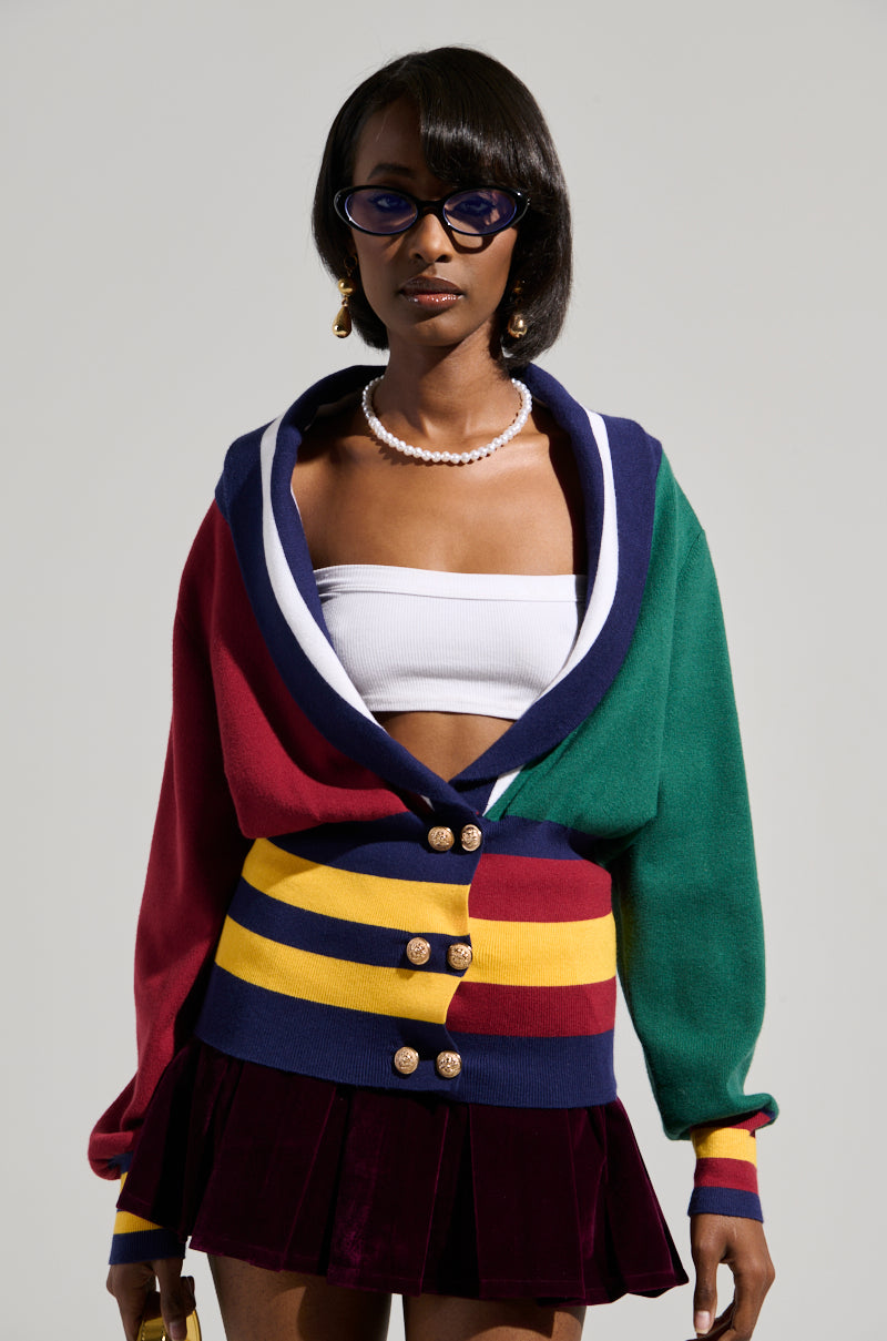 A person wearing the vibrant BOARDWALK BADDIE V-NECK STRIPED SWEATER with faux gold metallic closures, paired with a white strapless top and a burgundy skirt. They have short hair, sunglasses, and a pearl necklace, standing against a neutral background.