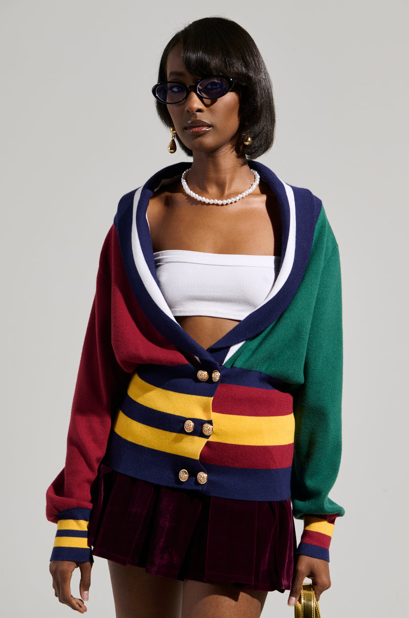 A person is stylishly dressed in the BOARDWALK BADDIE V-NECK STRIPED SWEATER, showcasing hues of green, red, blue, and yellow over a white top and a purple skirt. The ensemble features a color block pattern. They complement the look with short hair, sunglasses, a pearl necklace, faux gold metallic closures on the sweater, and gold earrings.