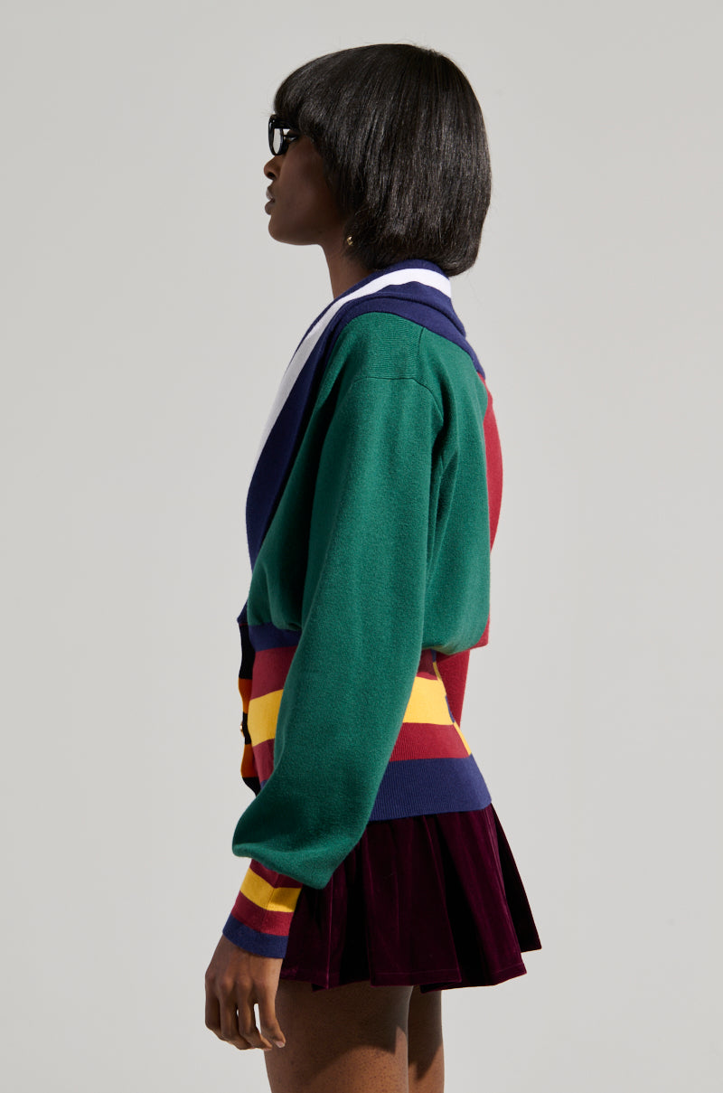 Wearing the BOARDWALK BADDIE V-NECK STRIPED SWEATER, a person with short dark hair showcases a vibrant color block pattern of green, blue, red, yellow, and white stripes. Paired with sunglasses and a burgundy pleated skirt to enhance the look, they stand in profile against a plain background.
