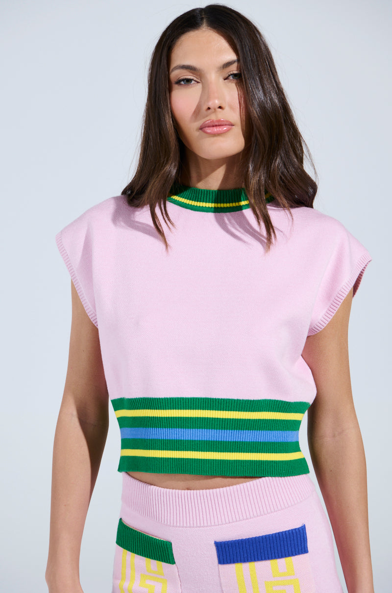A person with long dark hair is sporting the LOVE WINS SWEATER, featuring a sleeveless pink top with green and blue striped trim, paired with a matching skirt showcasing colorful geometric patterns. The background is plain and light-colored.