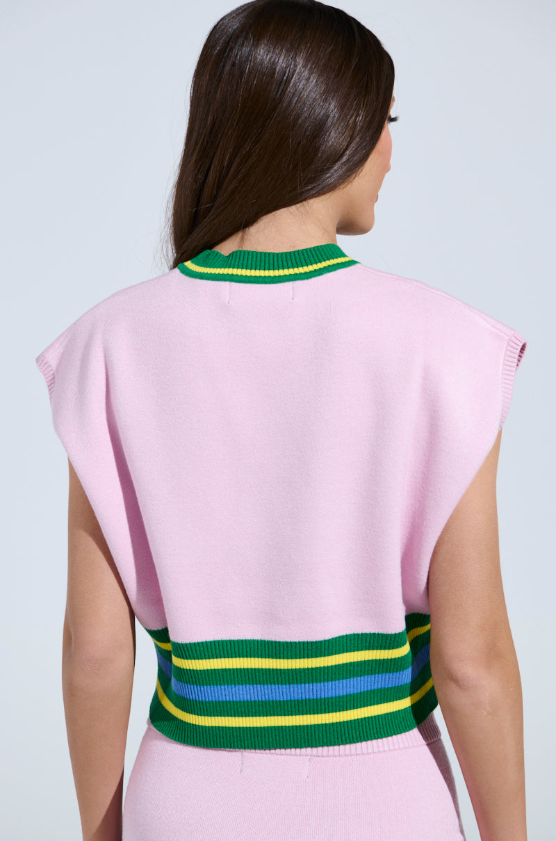 A person with long brown hair is seen from the back wearing the AKIRA Label's LOVE WINS SWEATER, a pink sleeveless top featuring green, blue, and yellow striped trim at the waist and neckline, embodying a sporty-chic style against a light gray background.