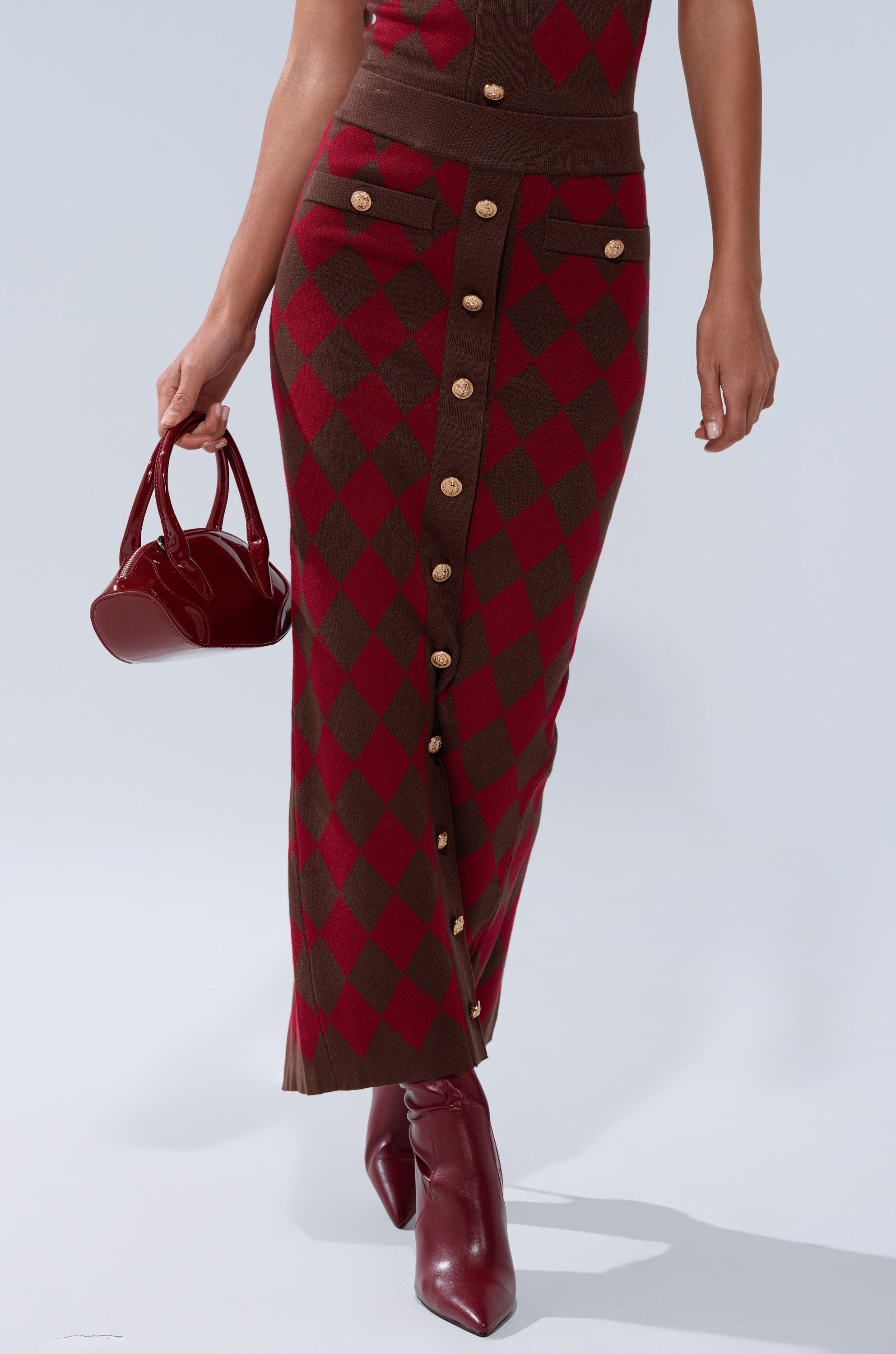 A person wearing THE BEST PART OF THIS ABSTRACT MAXI SKIRT with gold buttons, a matching top, maroon ankle boots, and carrying a small glossy maroon handbag.