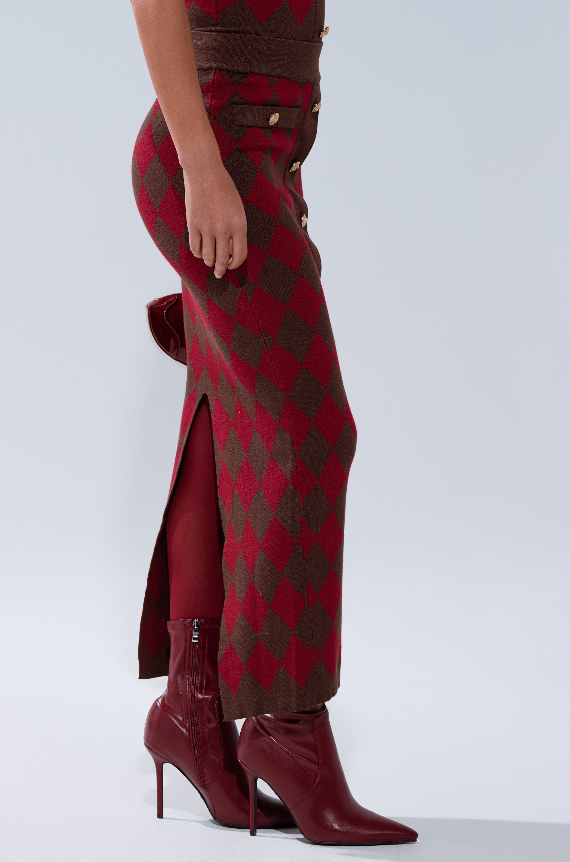 Wearing THE BEST PART OF THIS ABSTRACT MAXI SKIRT—a curve-hugging knit with red and brown diamond pattern, gold buttons, and a back slit—styled with red tights and pointed-toe burgundy ankle boots. Light gray background.