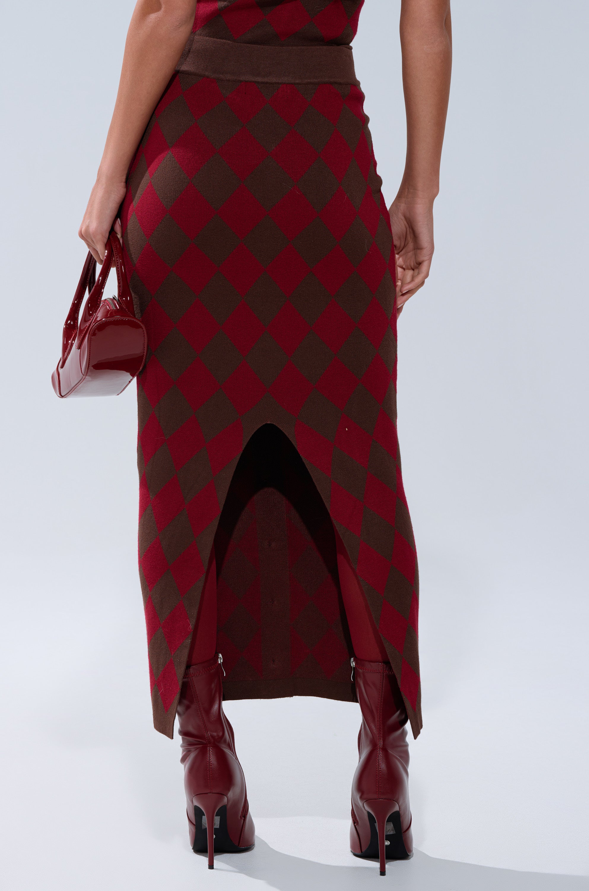 Photographed from behind, a person wears THE BEST PART OF THIS ABSTRACT MAXI SKIRT with a brown and red argyle pattern, featuring a back slit, paired with maroon high-heeled boots and a glossy red handbag.