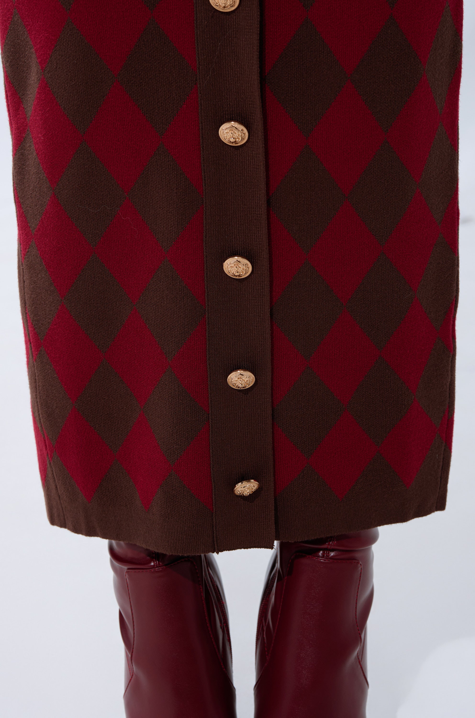 A close-up of THE BEST PART OF THIS ABSTRACT MAXI SKIRT, featuring a brown and red argyle pattern with gold center buttons, styled as a high-rise knit skirt, paired with shiny burgundy knee-high boots on a white background.