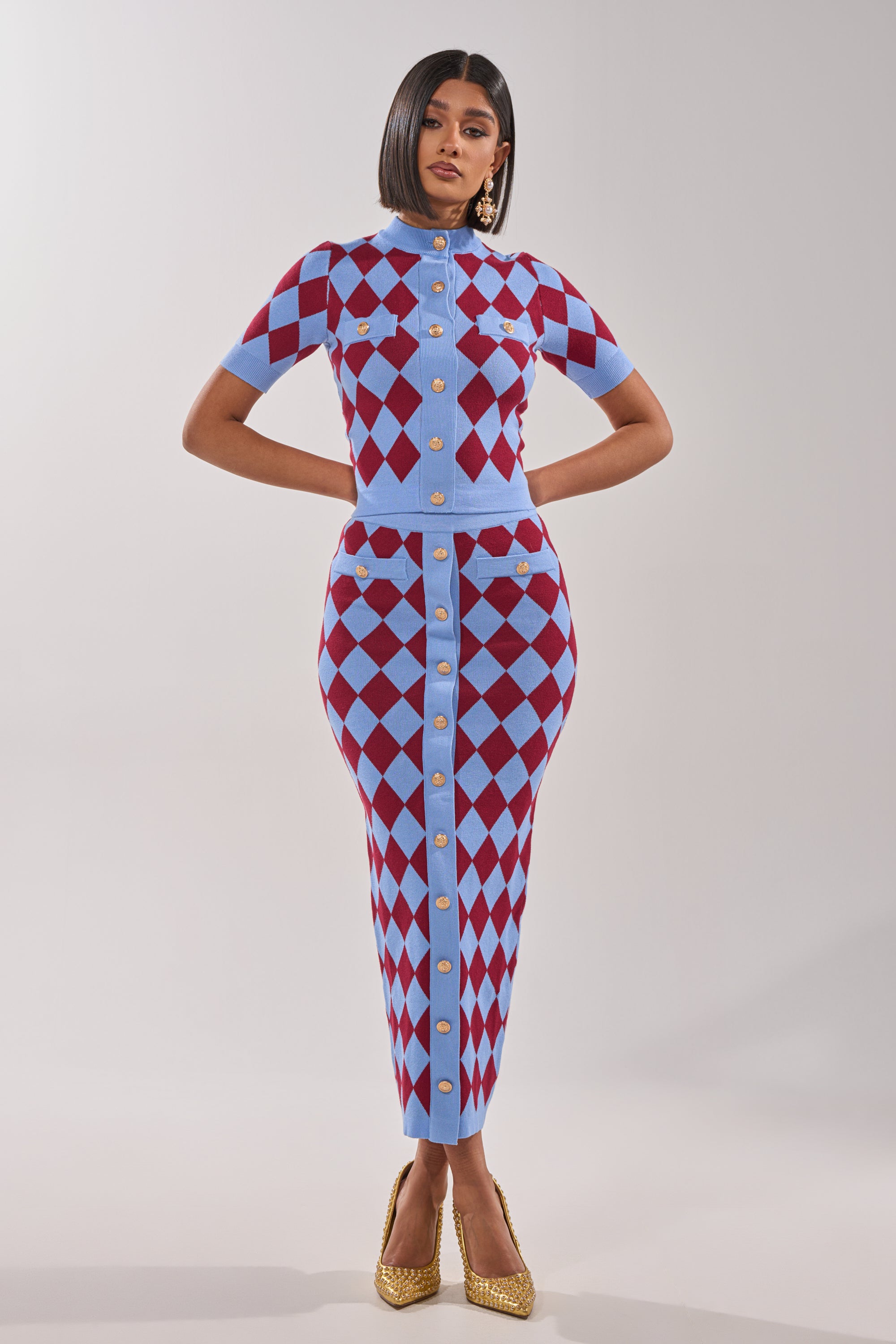 A woman stands confidently wearing the BABY GIRL SKIRT, a fitted short-sleeve dress featuring an argyle pattern skirt and gold buttons, paired with gold heels, large earrings, and a sleek bob hairstyle. 0