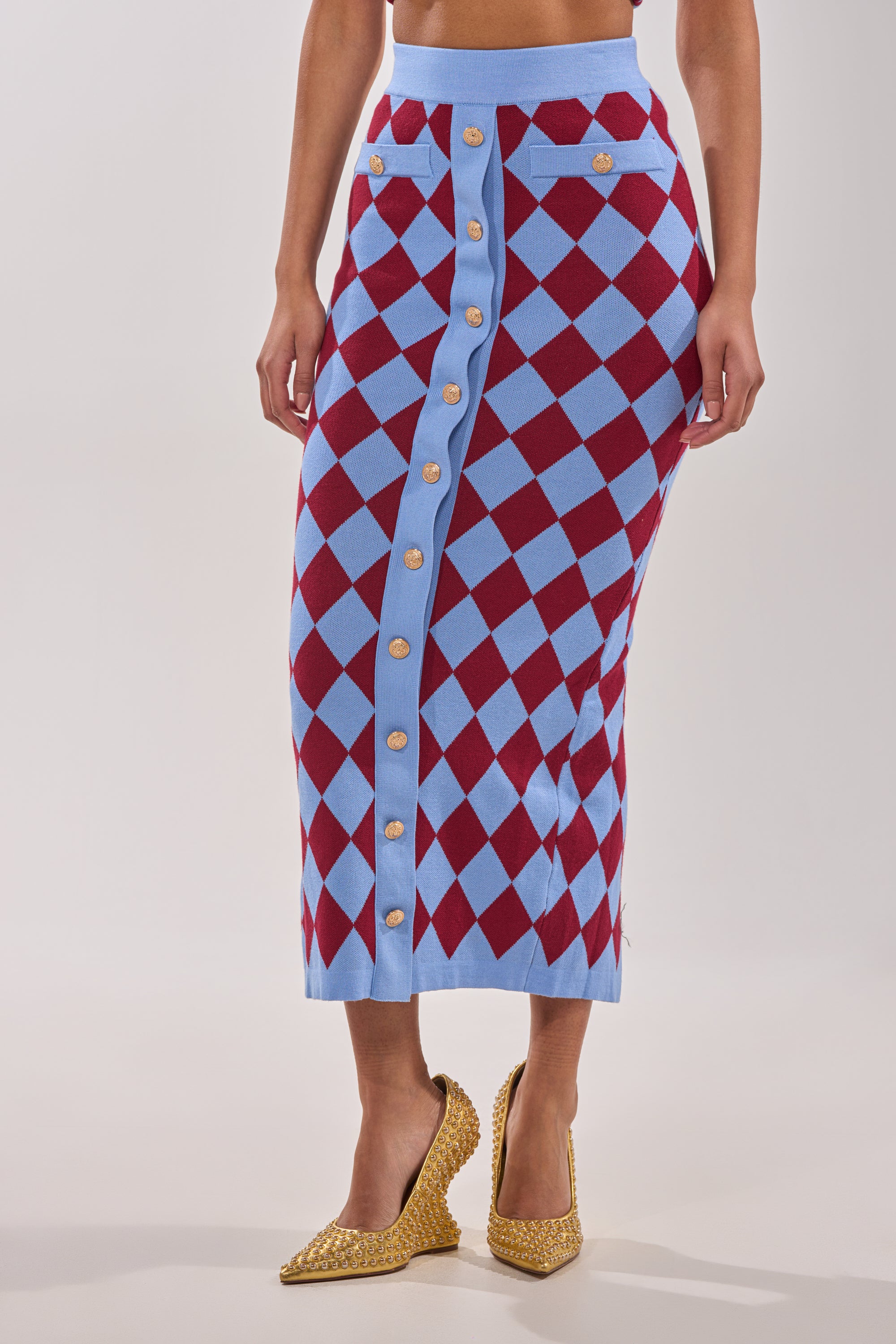 A woman wears the BABY GIRL SKIRT, showcasing an argyle pattern, high-rise waistband, and gold buttons down the front, styled with sparkly gold high-heeled shoes. 1