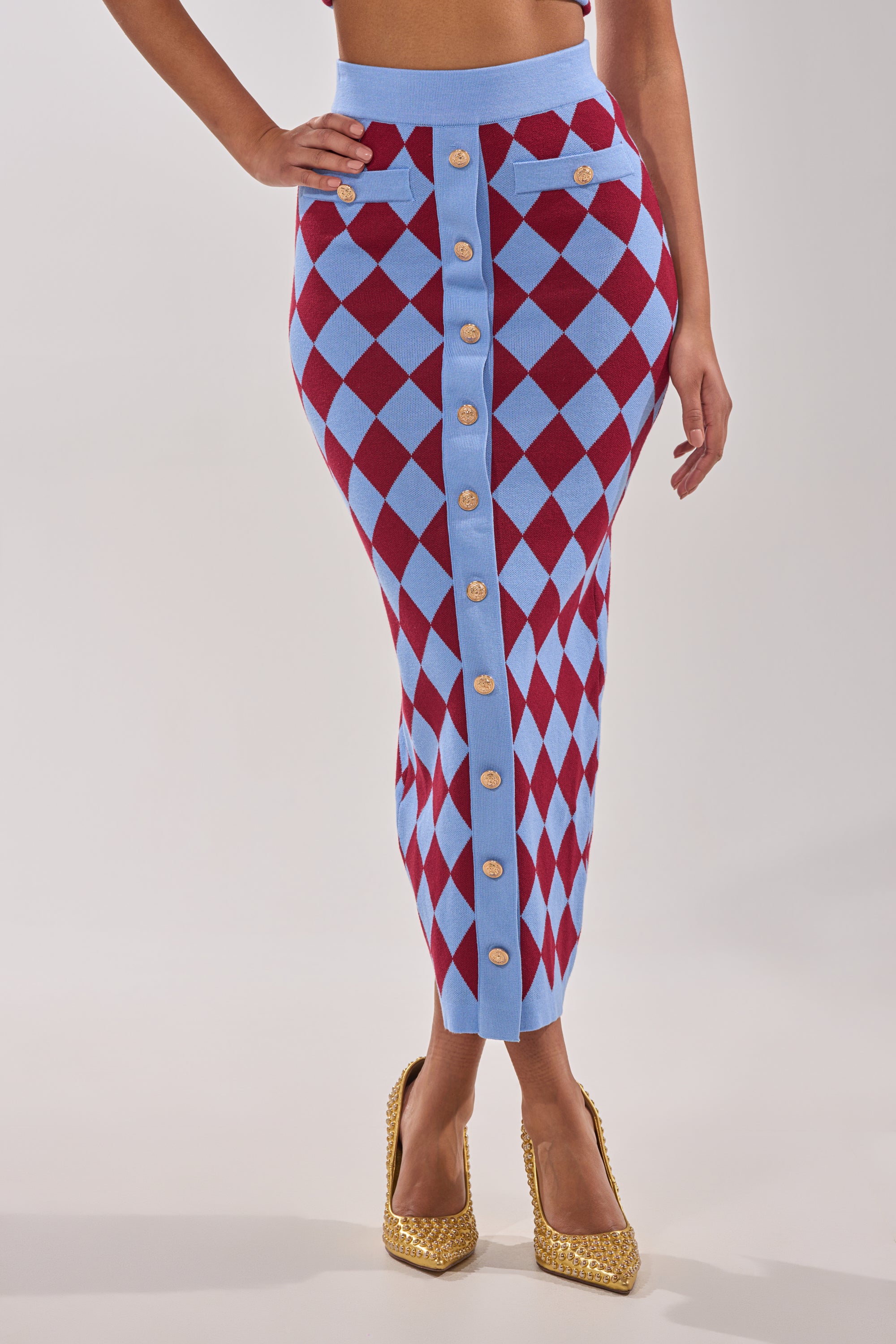 A woman stands against a white background, wearing the BABY GIRL SKIRT in blue and red argyle with gold buttons, styled with high-rise waistband and gold textured heels, one hand on her hip. 2