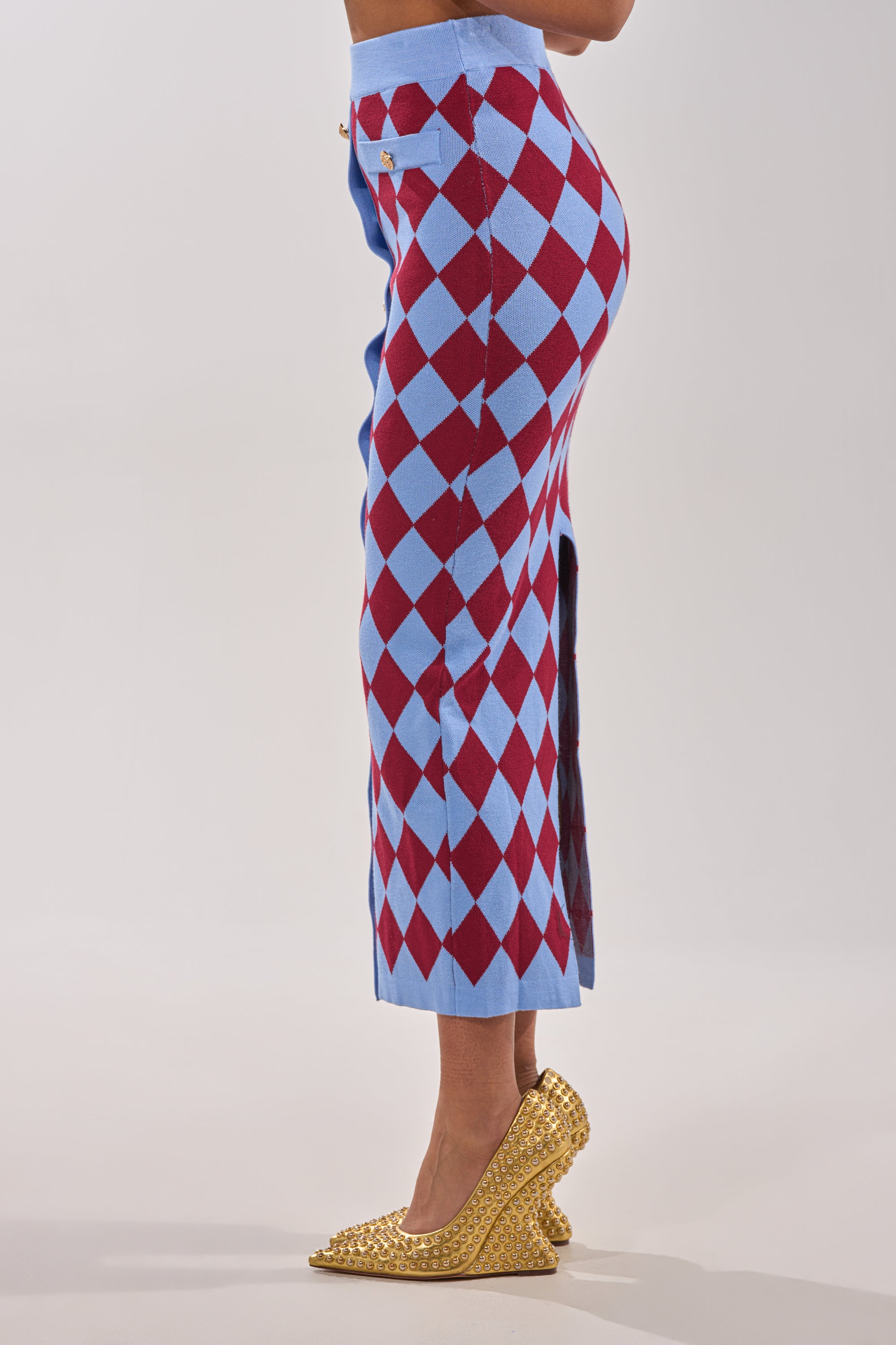 A woman stands sideways in the BABY GIRL SKIRT, a high-rise long skirt with blue and red argyle diamonds, a side slit, and gold studded high heels. The background is plain and light-colored. 3