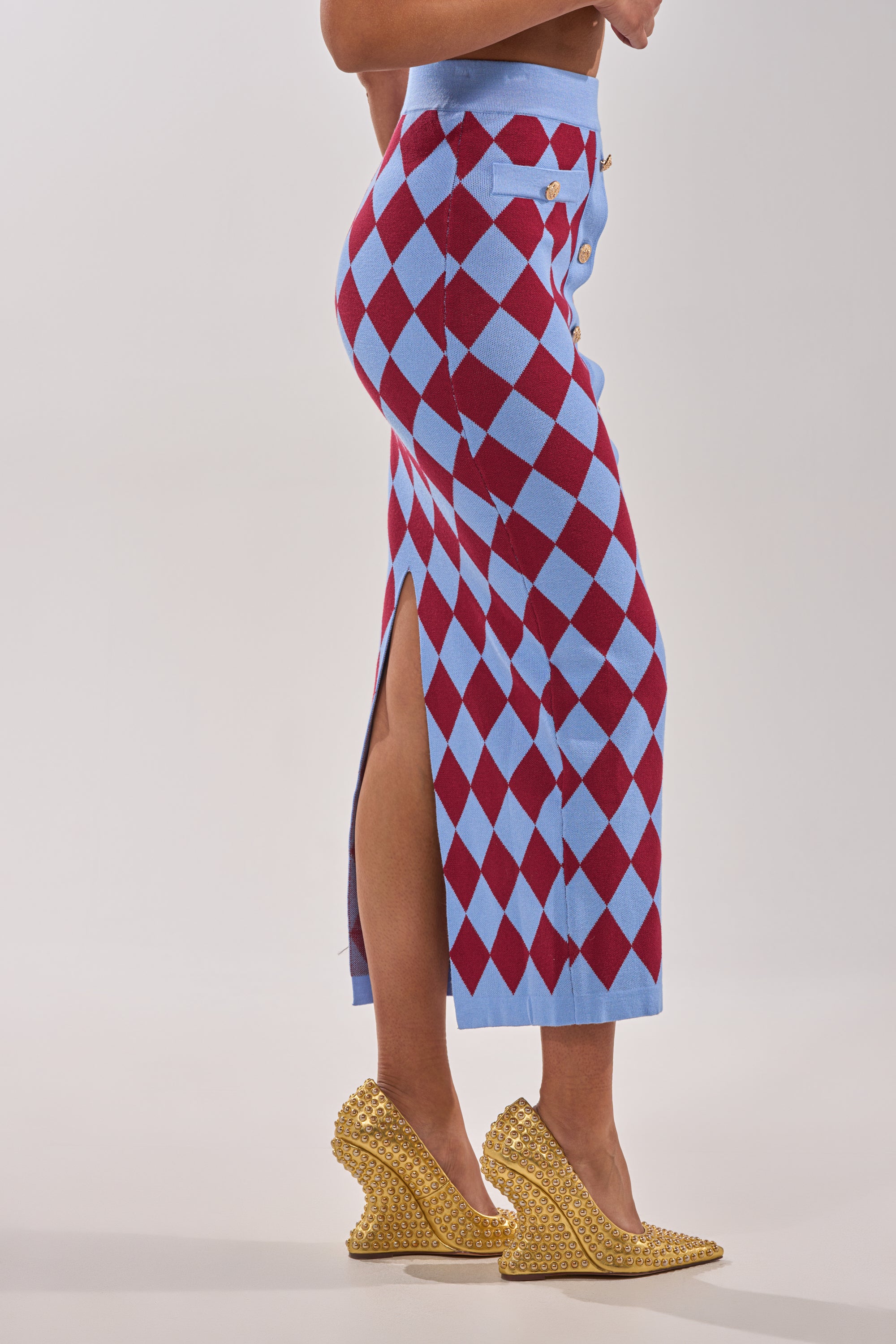 Wearing the BABY GIRL SKIRT featuring a high-rise waistband and red and blue argyle pattern with a side slit, paired with textured gold wedge heels, a person stands against a plain white background. 4