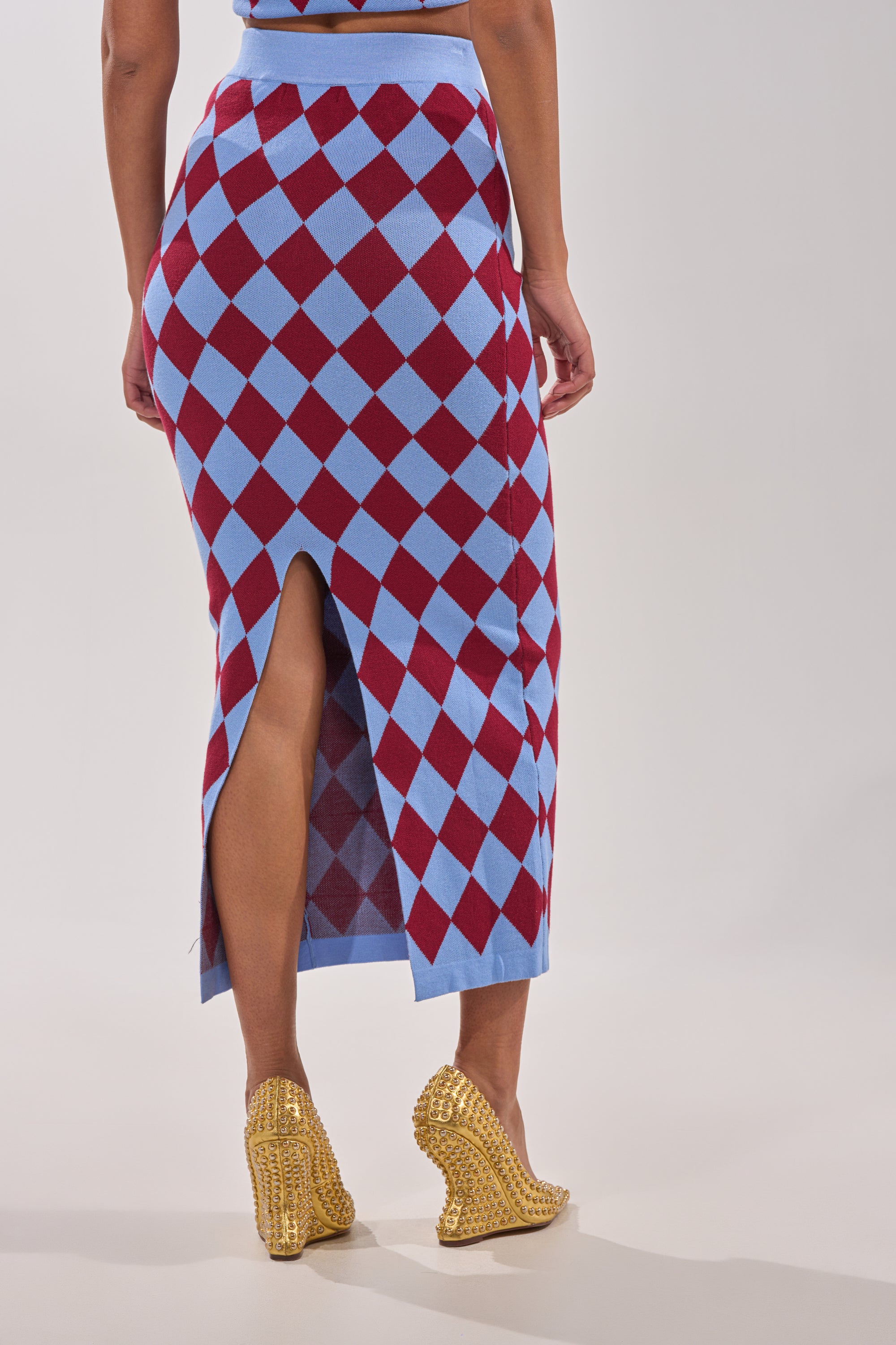 A person wears the BABY GIRL SKIRT, featuring a blue and red argyle pattern with a high-rise waistband and back slit. Paired with gold wedge heels, the cropped image shows the lower half of the body against a plain light background. 5