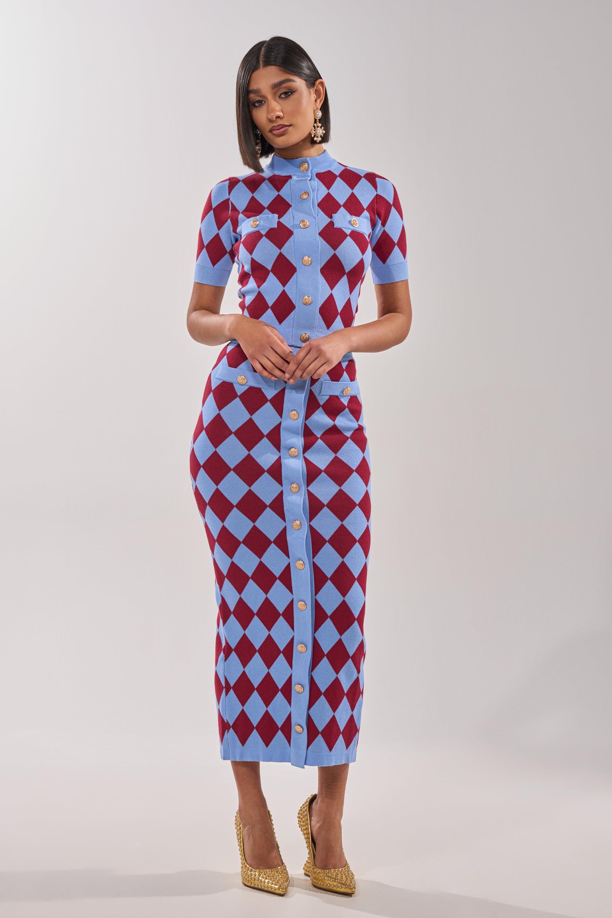 A woman faces forward in a fitted, short-sleeve dress featuring the BABY GIRL SKIRT with a blue and maroon argyle pattern, high-rise waistband, gold front buttons, and accessorizes with gold earrings and heels against a plain light background. 6