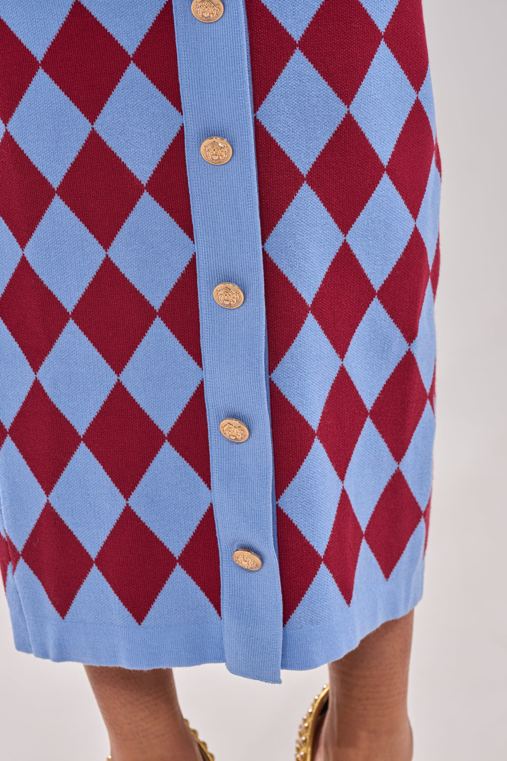 The image shows a close-up of the BABY GIRL SKIRT, featuring a blue and red argyle pattern, high-rise waistband, gold buttons down the front, and a visible hem along with part of the wearer’s legs and shoes. 7