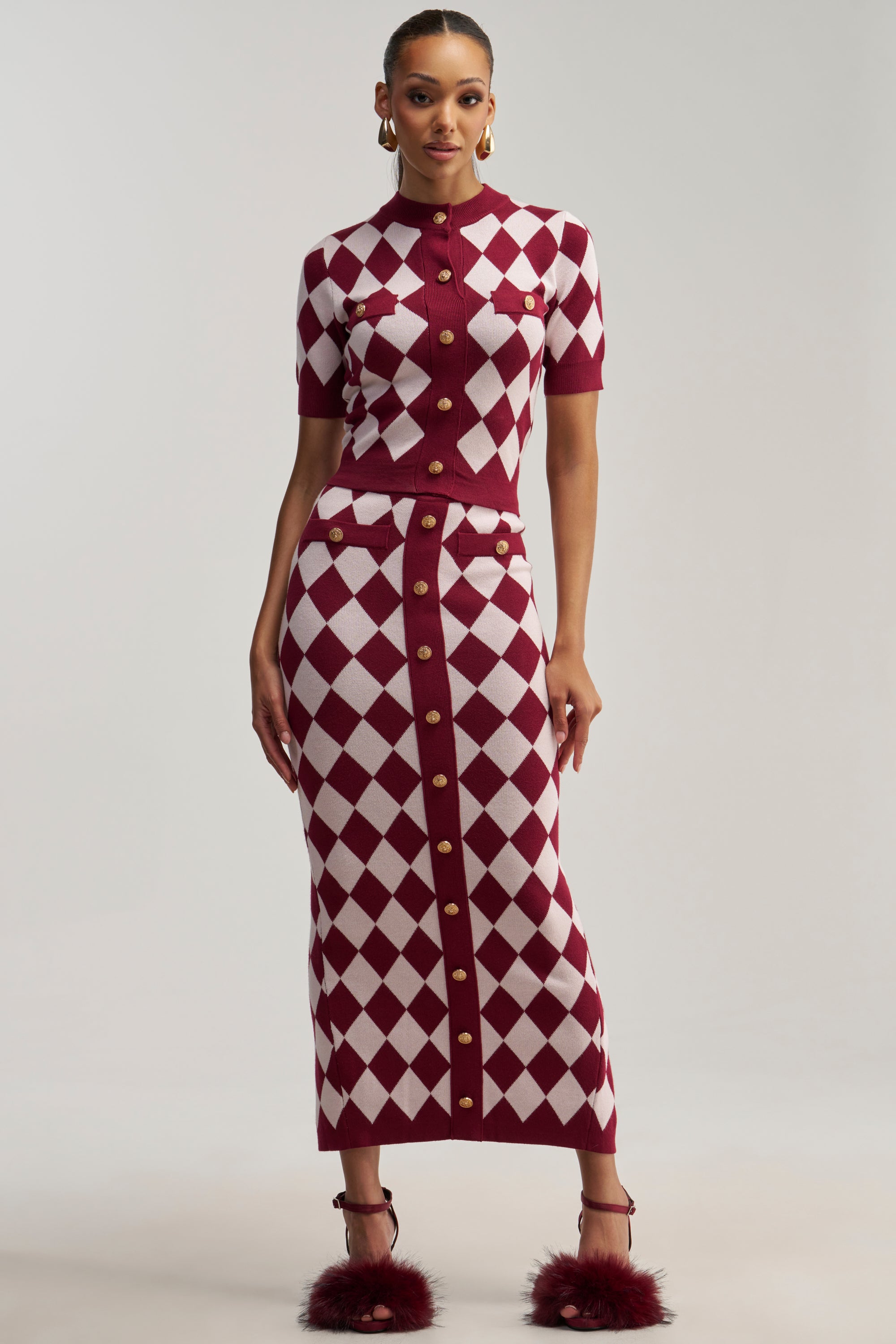 A woman wears a fitted, short-sleeve, maroon and white checkered knit top paired with the SWEET BABY GIRL MIDI SKIRT IN RED MULTI, which is a matching high-rise knit skirt with gold buttons. She completes the look with maroon feathered high-heeled sandals.