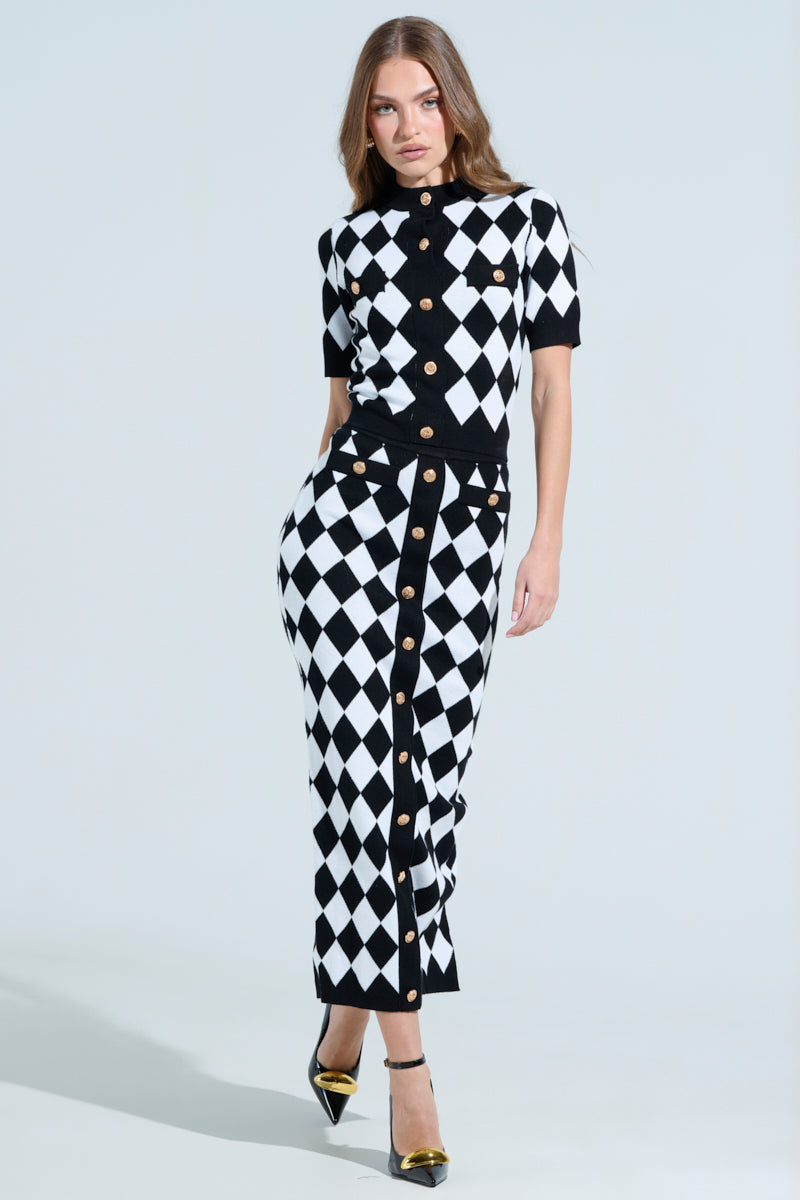 A woman flaunts a fitted black and white harlequin-patterned maxi skirt, accentuating a bodycon silhouette. She pairs it with pointed heels featuring metallic details, confidently standing against a plain background.